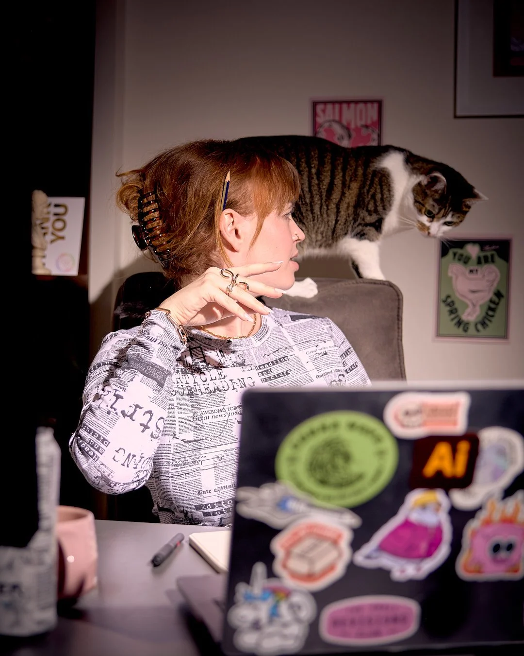 A woman sitting at a desk with a laptop covered in colorful stickers, a pen, a notebook, and a pink mug, with a tabby cat walking on her chair back, in a room with wall art.