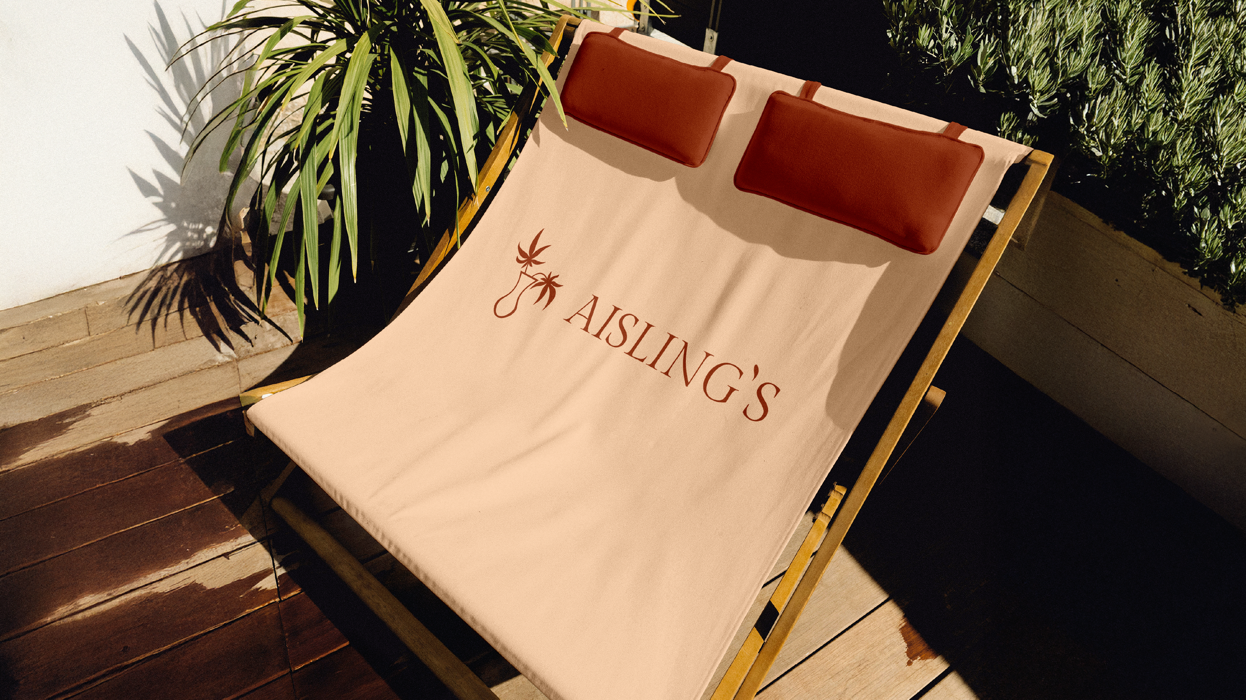 Outdoor lounge chair with a beige fabric cover, featuring a logo with a star and palm trees, and the words 'Aisling's' written underneath in red, positioned on a wooden deck with plants nearby and a white wall in the background.