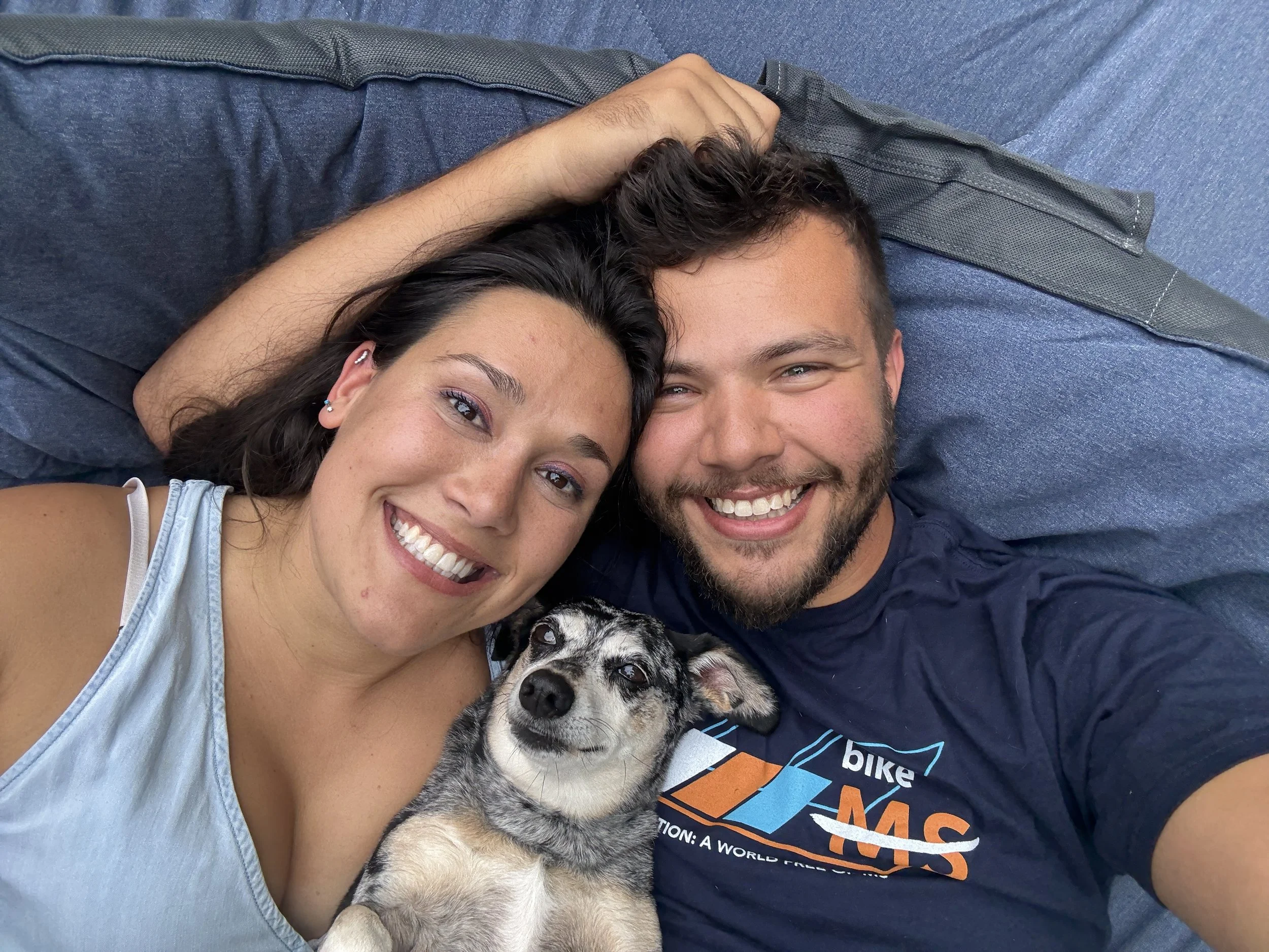 A smiling couple lying on a blue couch with their dog, a Siberian Husky mix, between them.