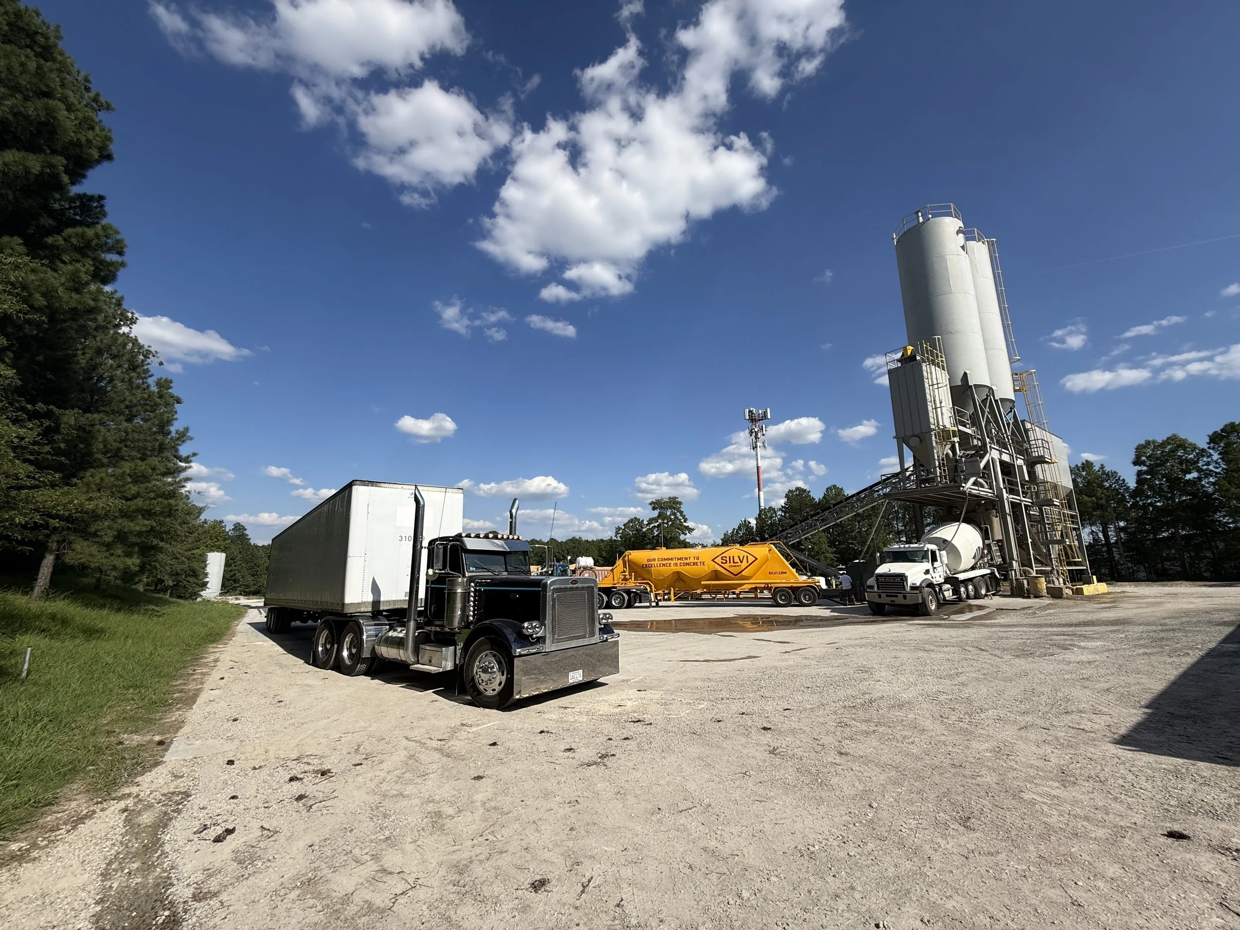 A construction site with a cement silo, trucks, and a yellow concrete mixer truck labeled 'SILVI', surrounded by trees under a blue sky with white clouds.