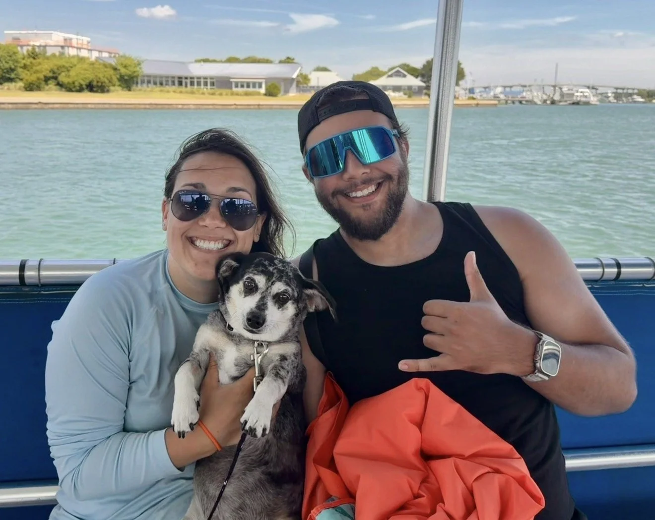 Smiling woman and man on boat holding a dog, water and a bridge in the background.