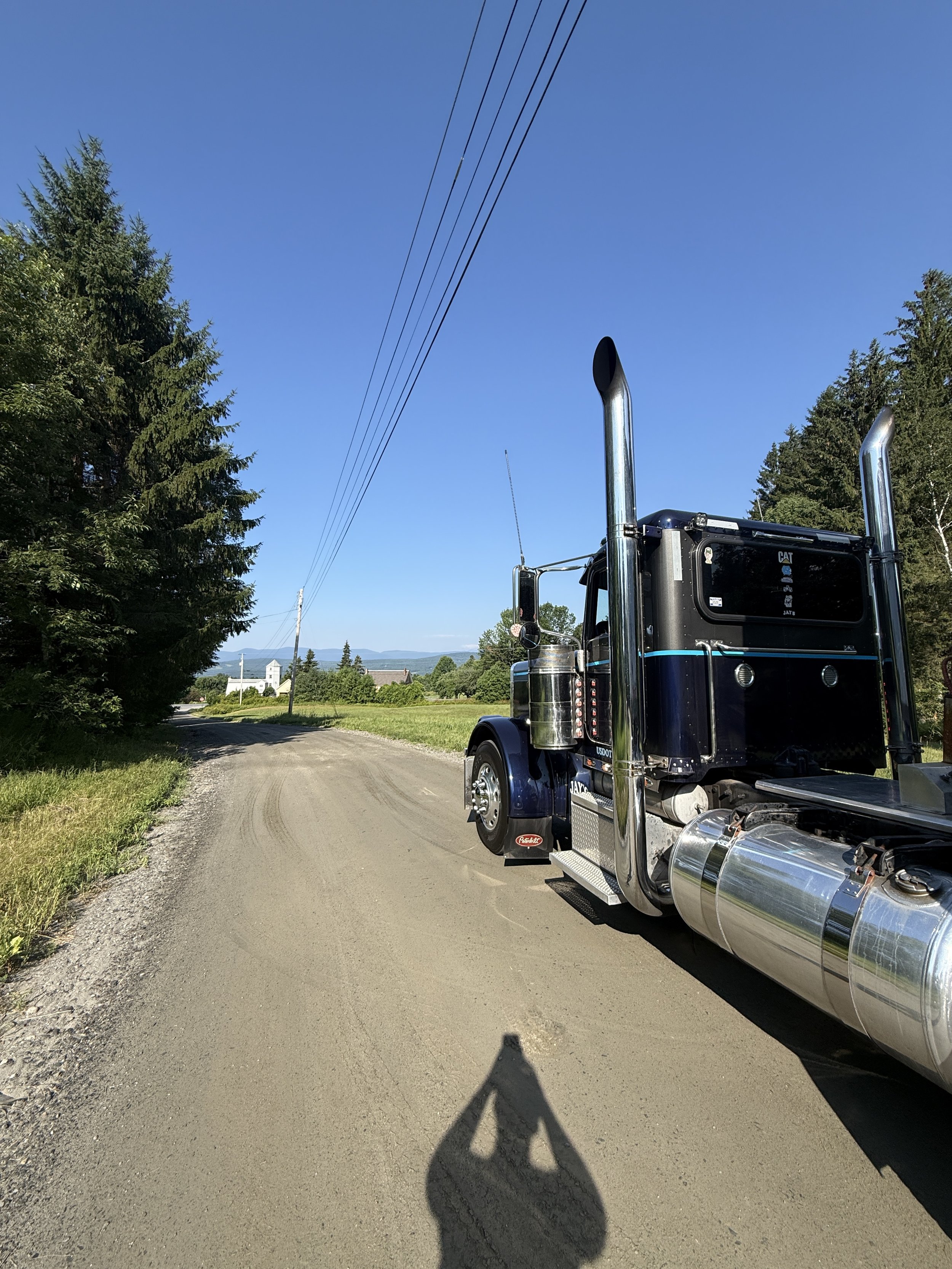A black semi-truck parked on a dirt road with trees on both sides and a blue sky overhead.
