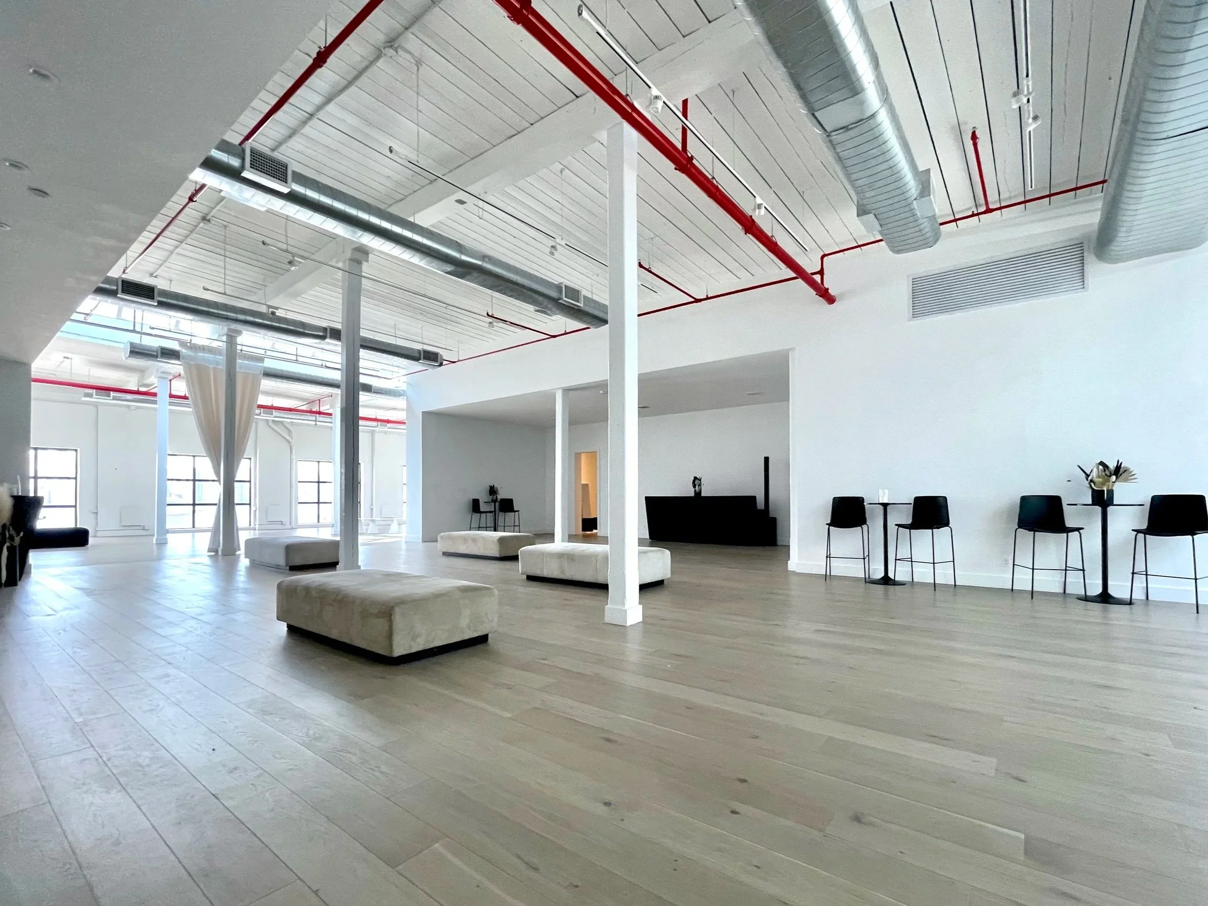 The Brooklyn Rare Book Fair venue: A spacious, modern indoor lounge with white walls, a light wooden floor, large windows, and various seating options including a white ottoman and black chairs.