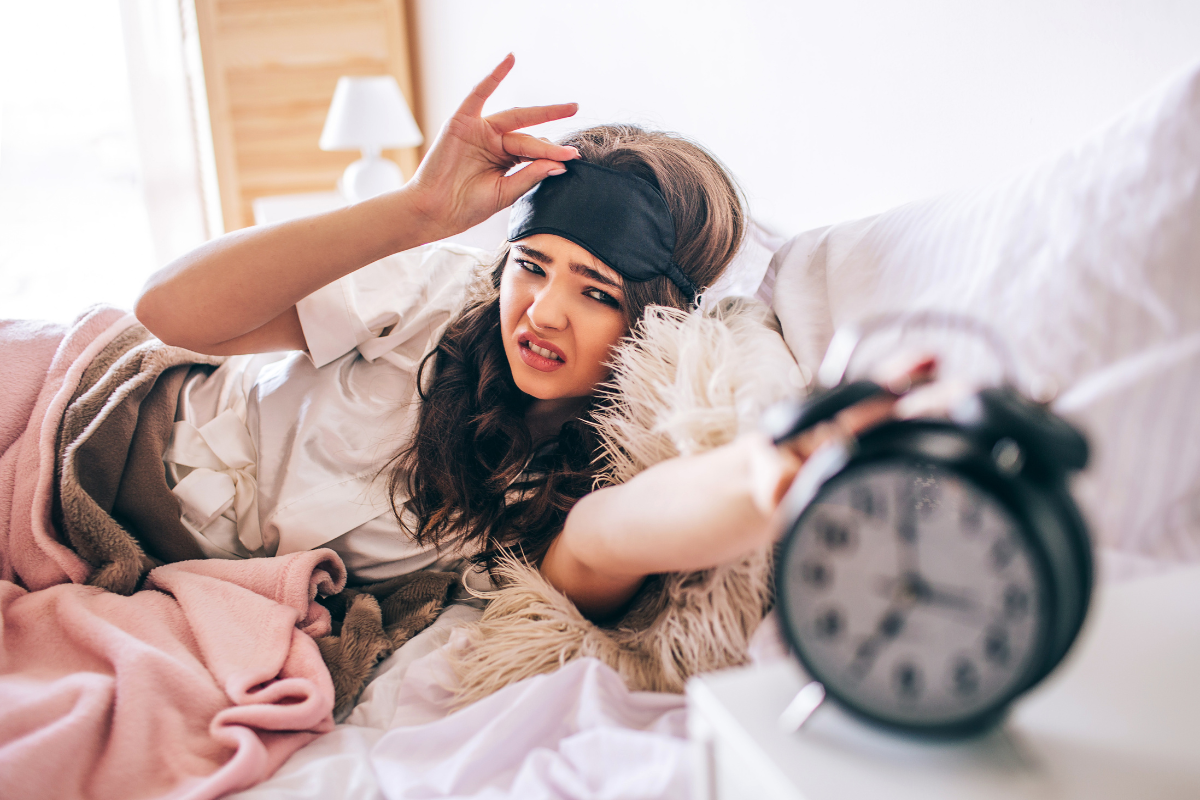 A woman lying in bed with a headache, holding an ice pack to her forehead, with an alarm clock showing 7:50 nearby.