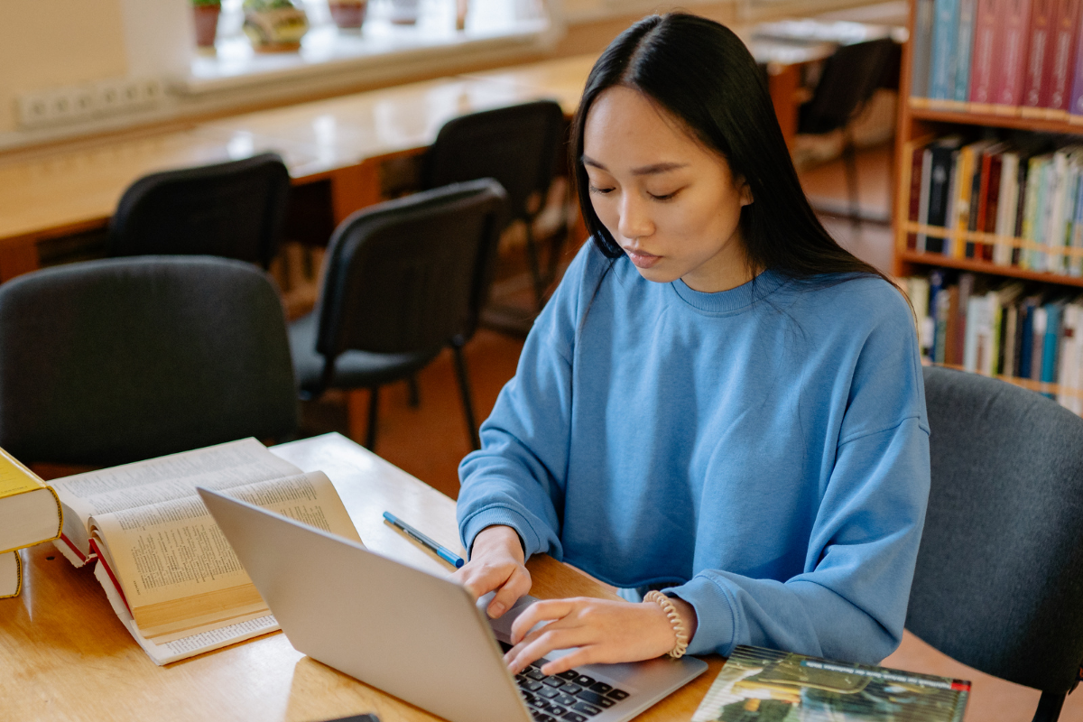 A young woman with long black hair wearing a blue sweatshirt sitting at a wooden table in a library, working on a laptop, with books and a pen nearby.