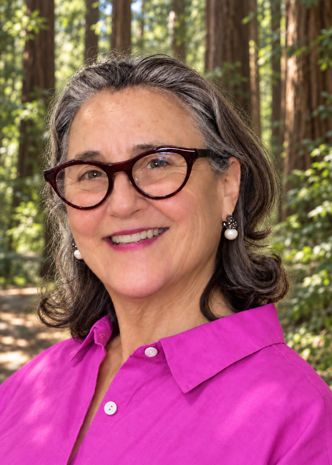 A woman, Coach Julie, with gray hair, glasses, and pearl earrings smiling outdoors in a forest setting, wearing a purple shirt.