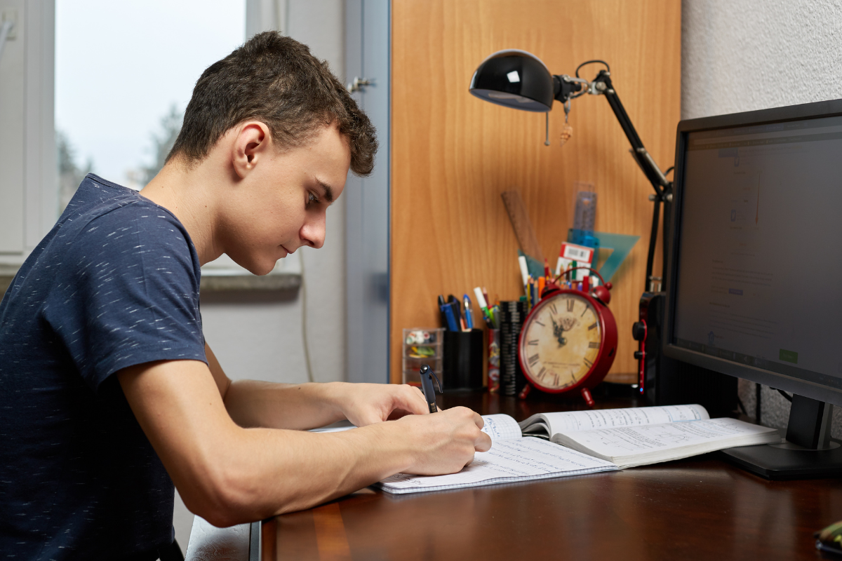 Young man studying and writing notes at a desk with a computer, clock, and office supplies nearby.