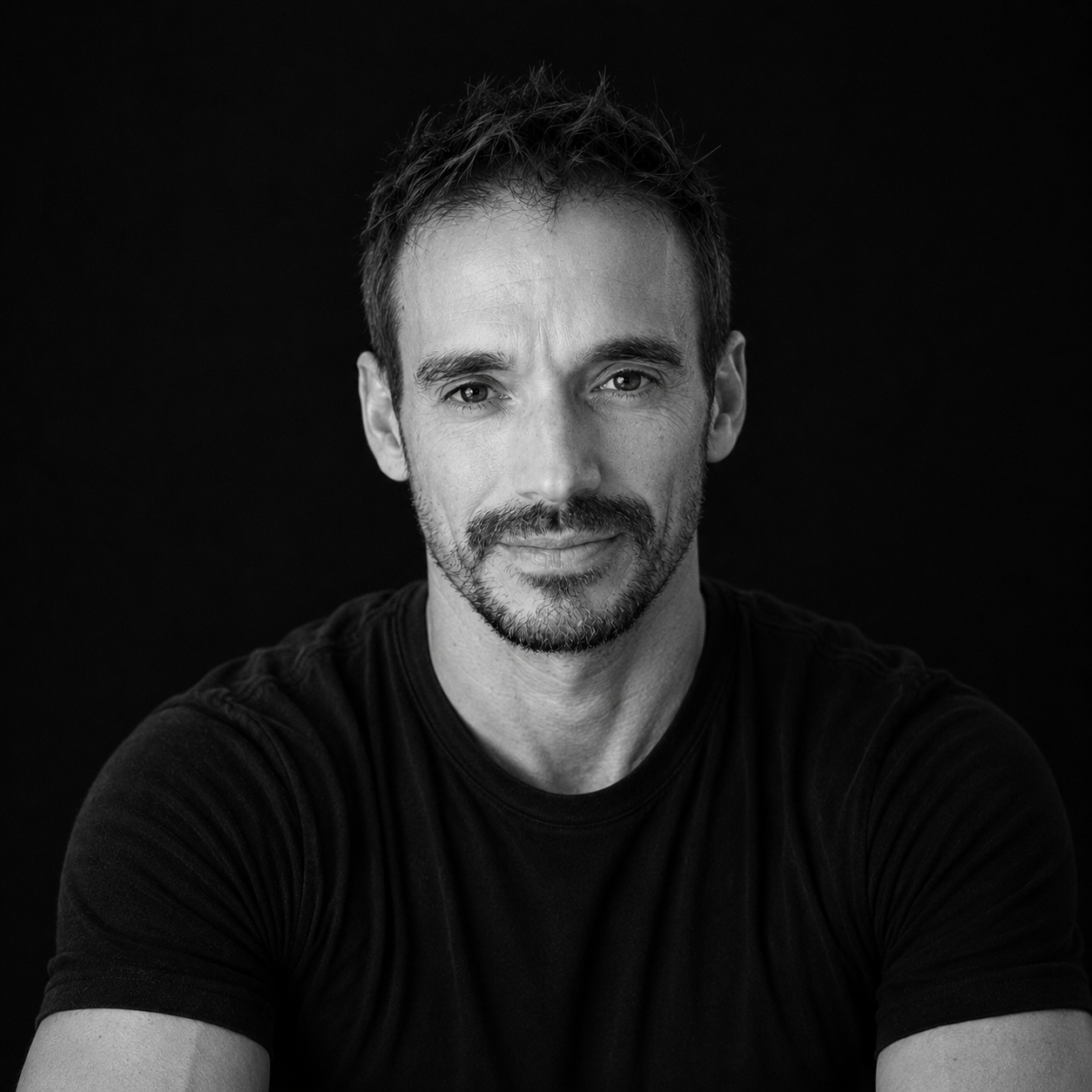 Black and white portrait of Coach Rod with short, tousled hair, facial hair, and a slight smile, wearing a black t-shirt against a dark background.
