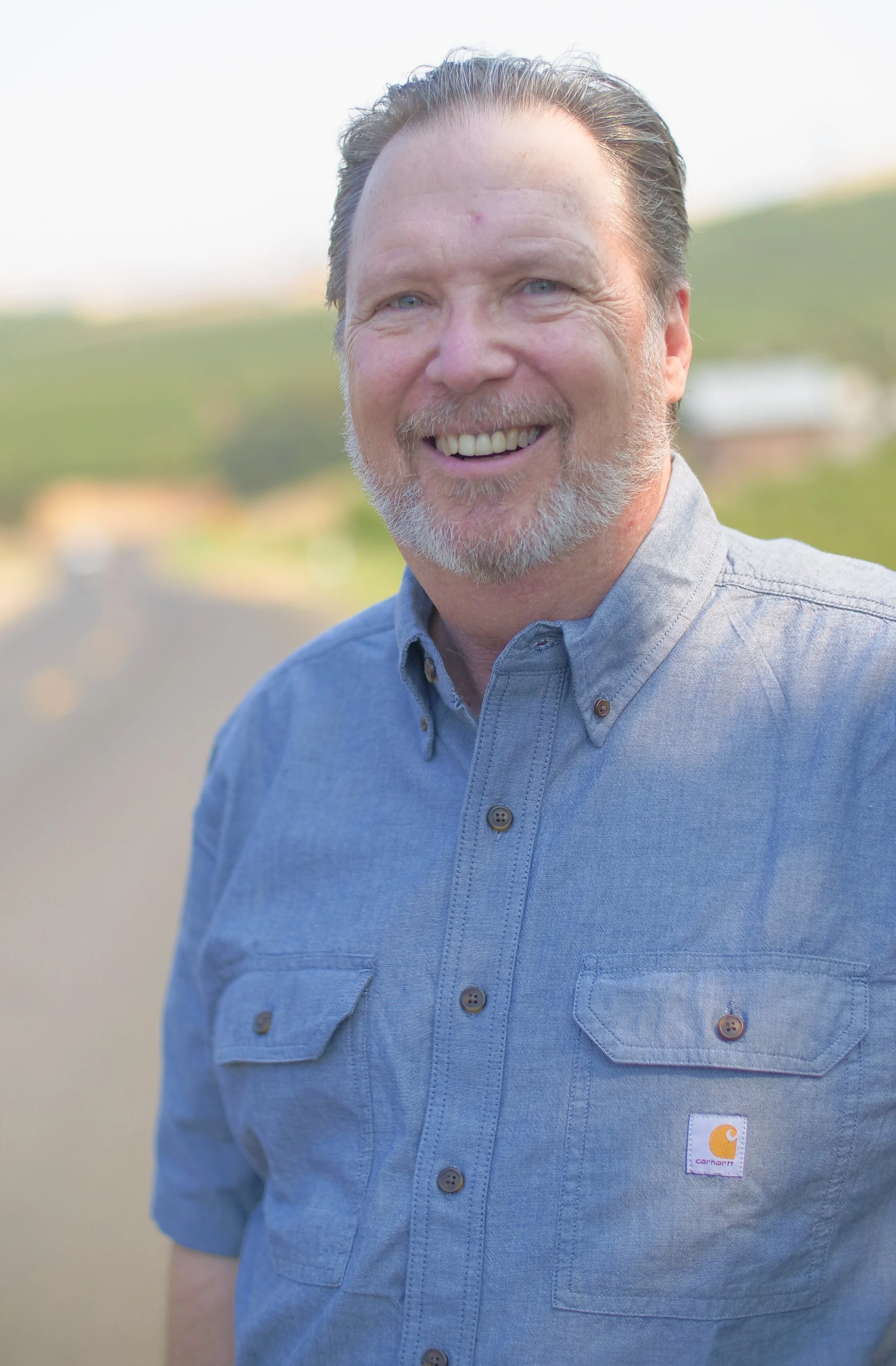 A smiling man with a beard and blue eyes, wearing a light blue Carhartt shirt, standing outdoors with a blurred background of greenery and sky.