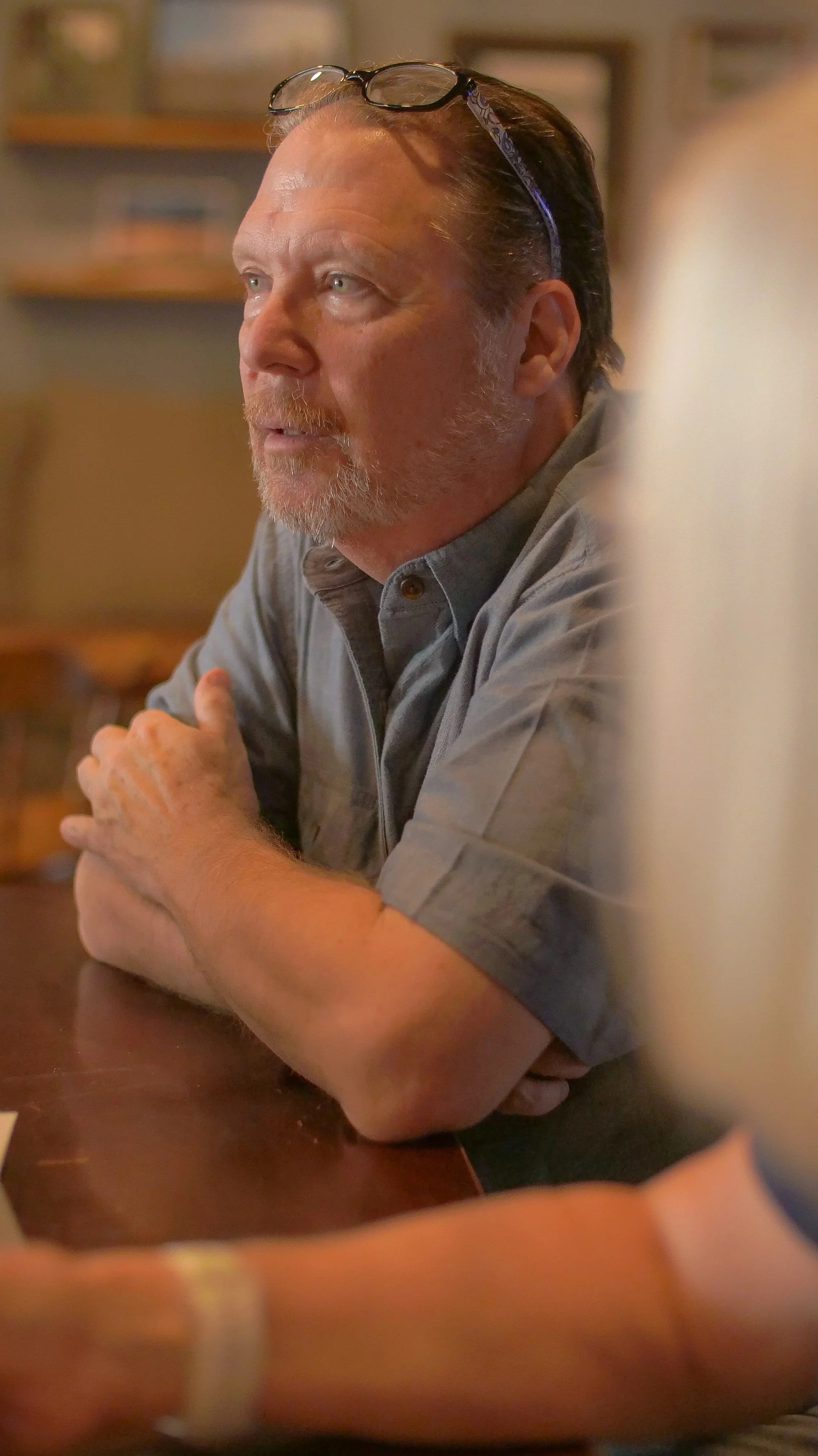 A middle-aged man with glasses on his head, a beard, and a mustache, leaning on a wooden table with his arms crossed, in a relaxed setting.