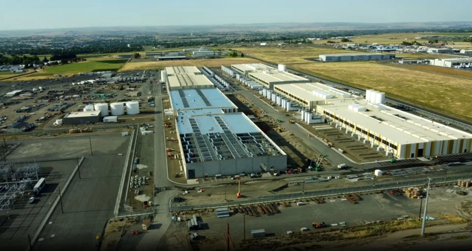 Aerial view of an industrial complex under construction with multiple buildings, parking lots, and storage tanks, situated in a rural area with open fields in the background.