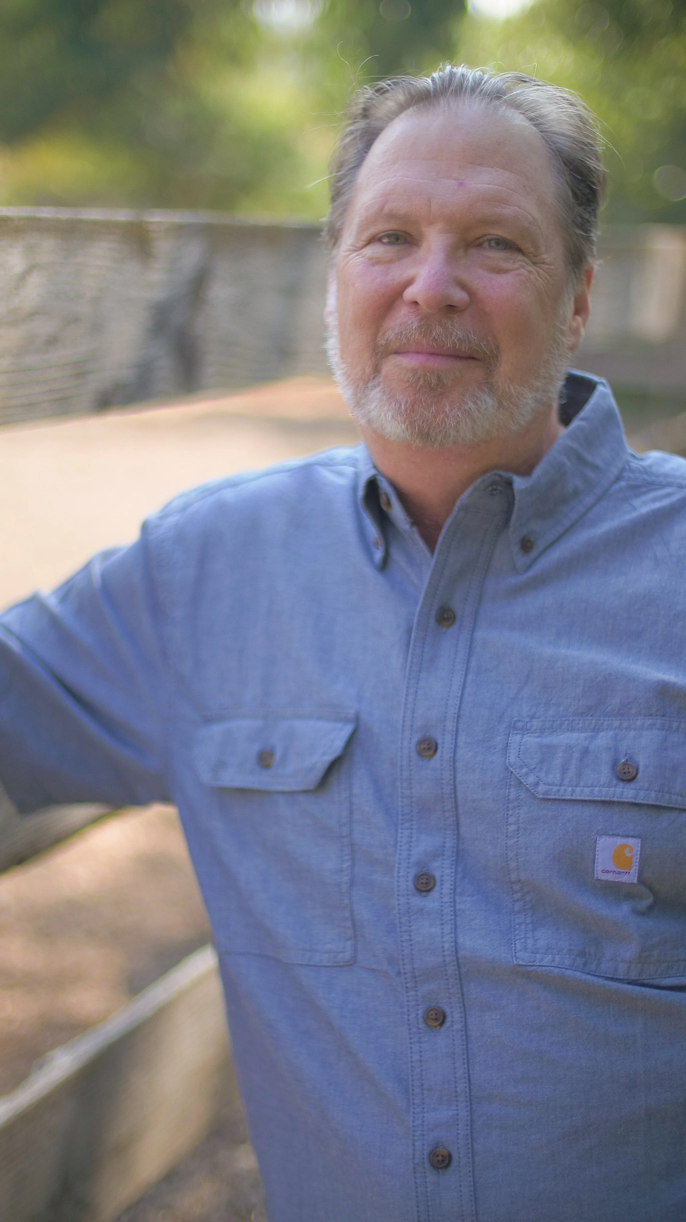Close-up of a middle-aged man with gray hair, a beard, and light blue eyes. He is wearing a light blue button-down shirt and standing outdoors in a park-like setting with blurred green trees and fencing in the background.