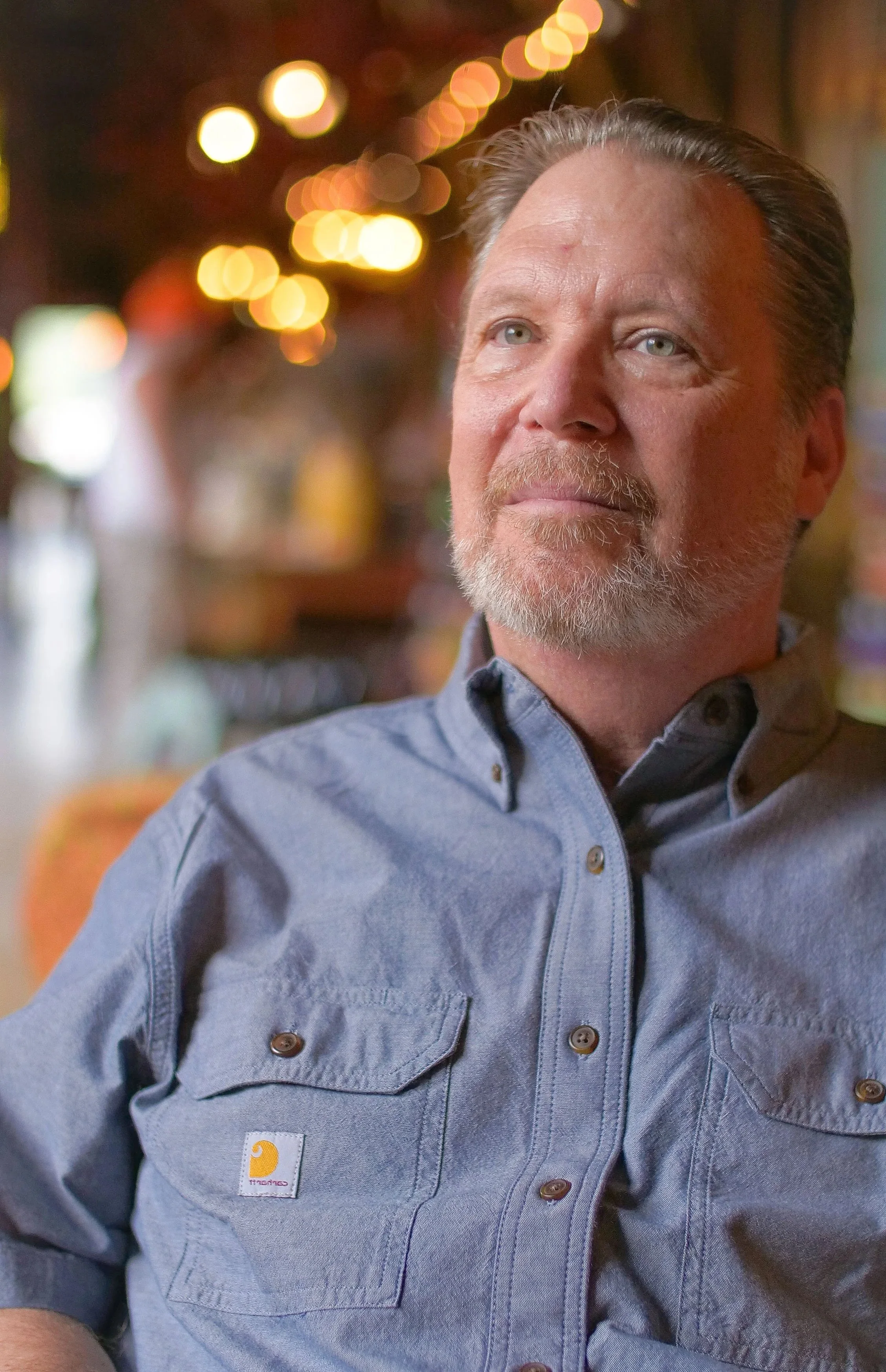 A middle-aged man with gray hair and beard, wearing a light blue Carhartt shirt, sitting indoors with blurred background of warm lighting.