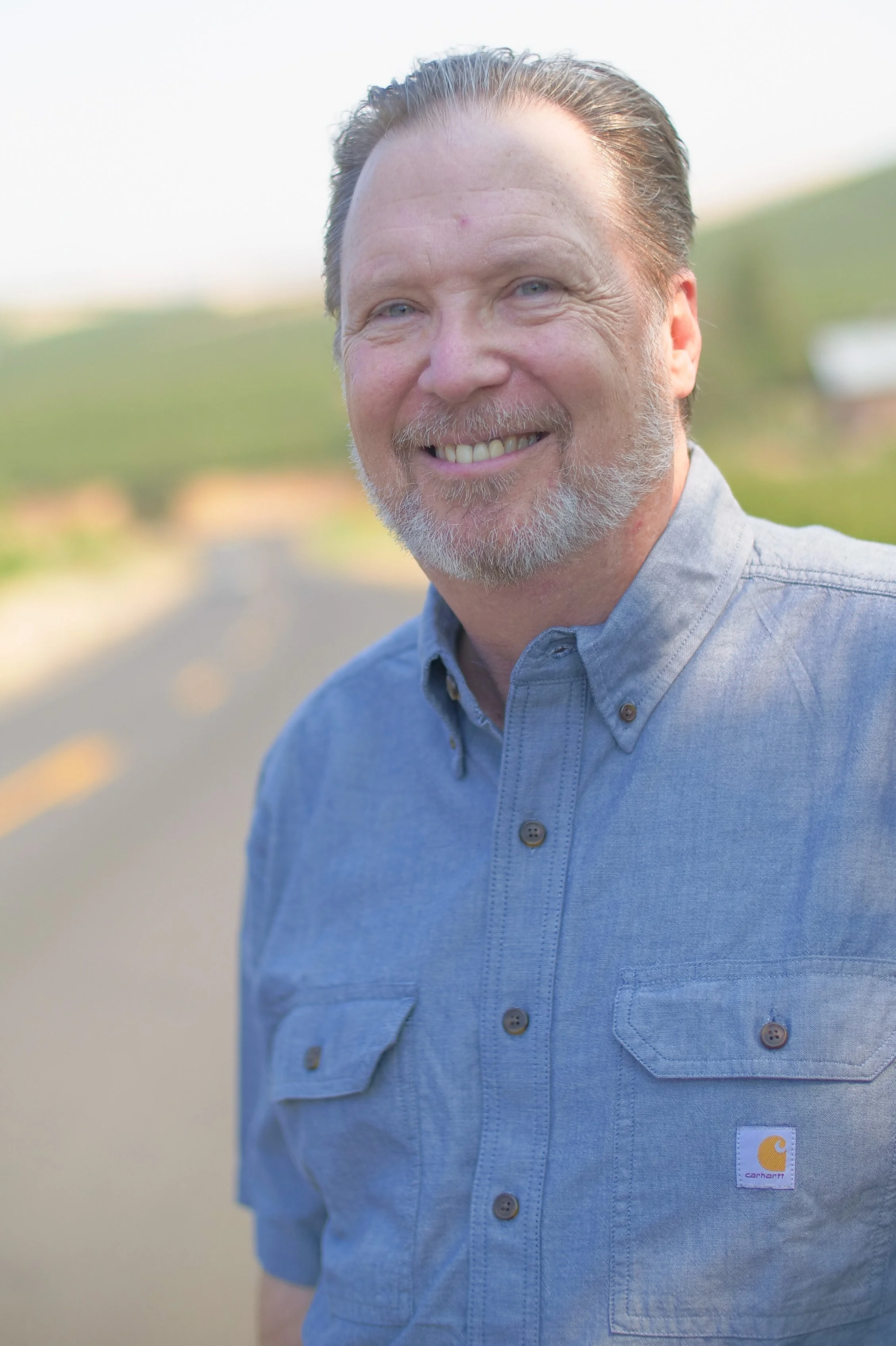 A middle-aged man with light skin, short gray and white hair, and a beard, smiling outdoors. He is wearing a light blue Carhartt shirt with a logo tag, standing on a paved road with a blurred background of green fields and a partly cloudy sky.