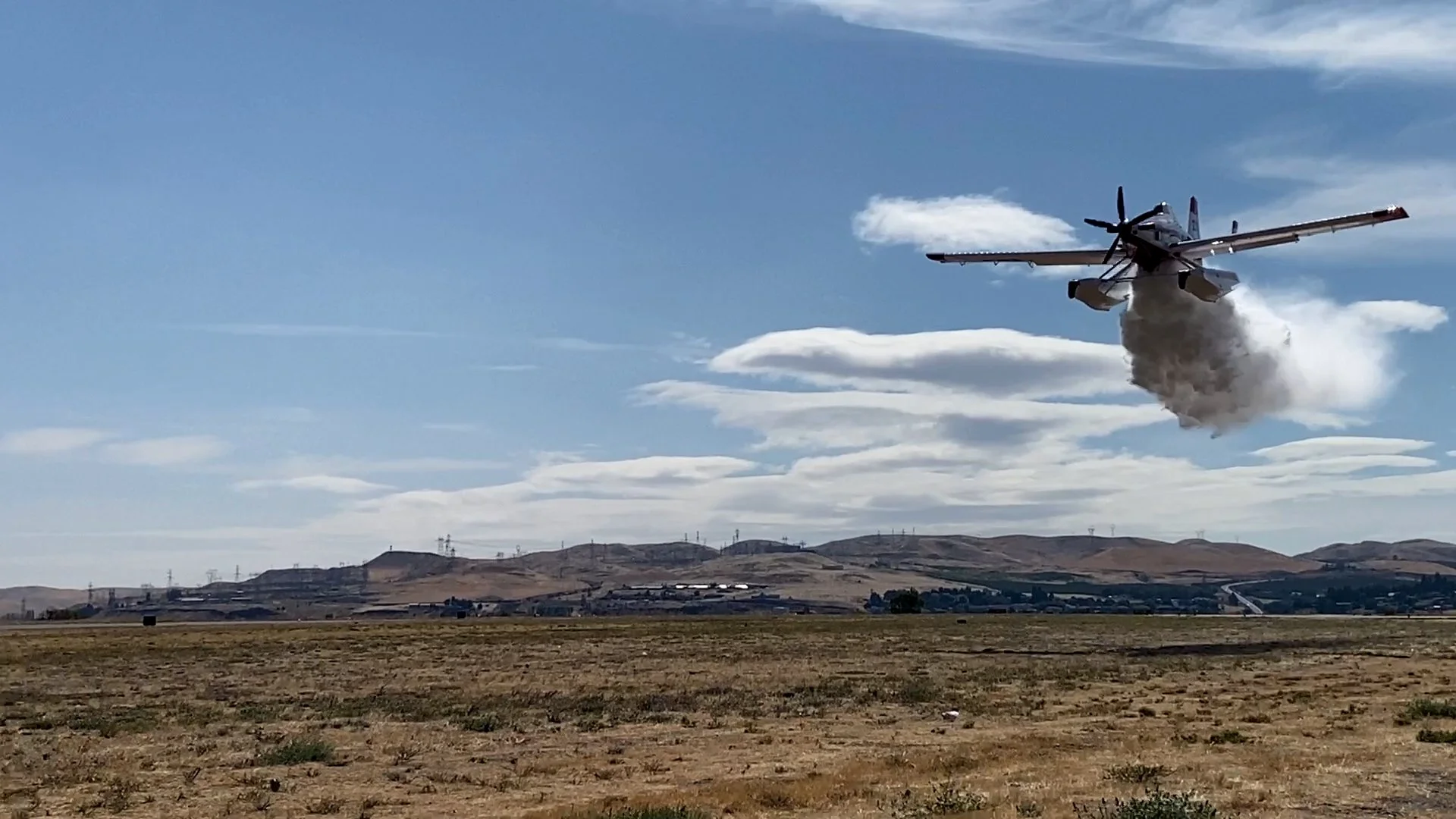 A fighter jet airplane is taking off, leaving a trail of dust and smoke behind. The landscape is dry with hills in the distance, and the sky is partly cloudy.