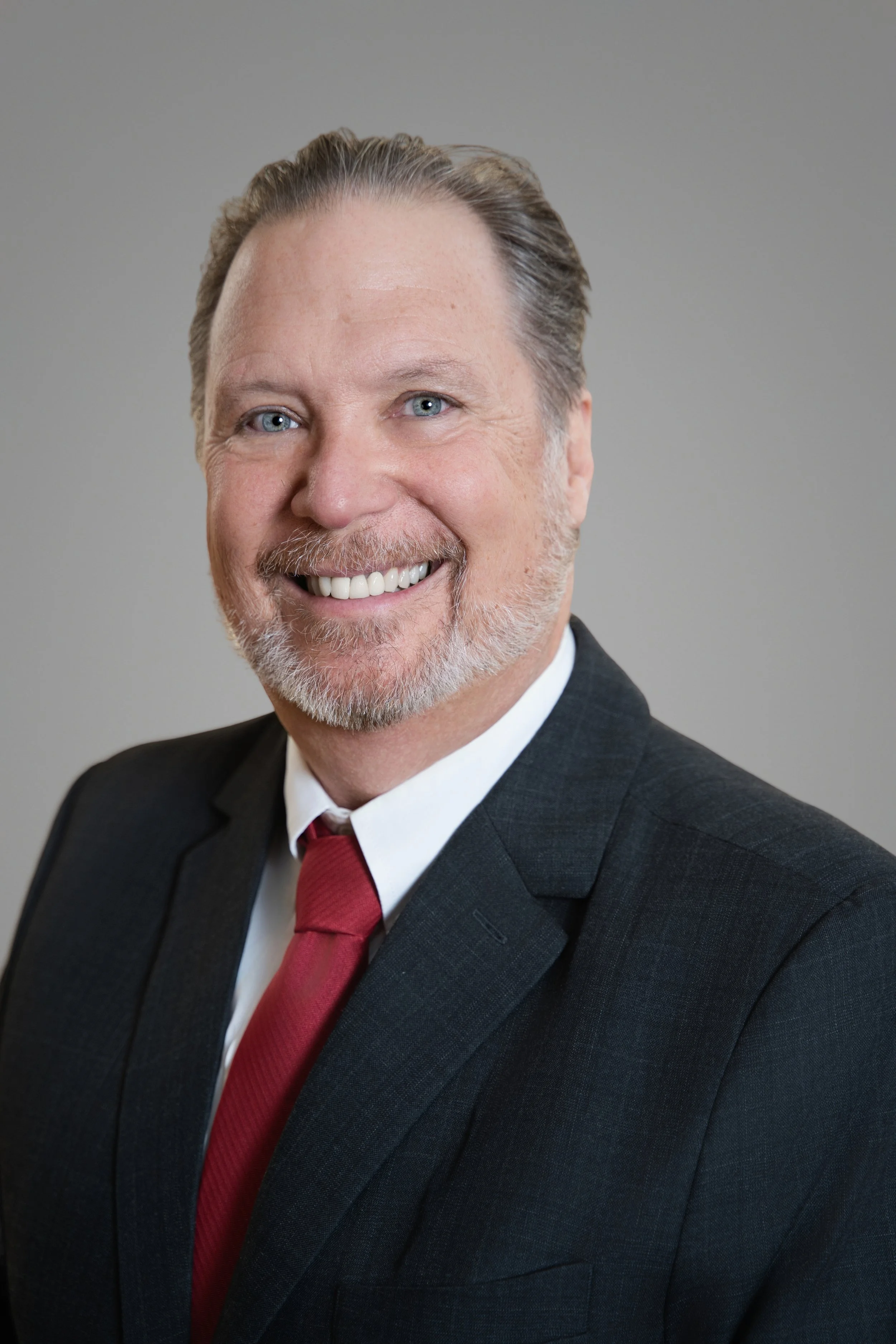Close-up of a smiling middle-aged man with blue eyes, gray hair, and a beard, dressed in a dark pinstripe suit, white shirt, and red tie, against a plain gray background.