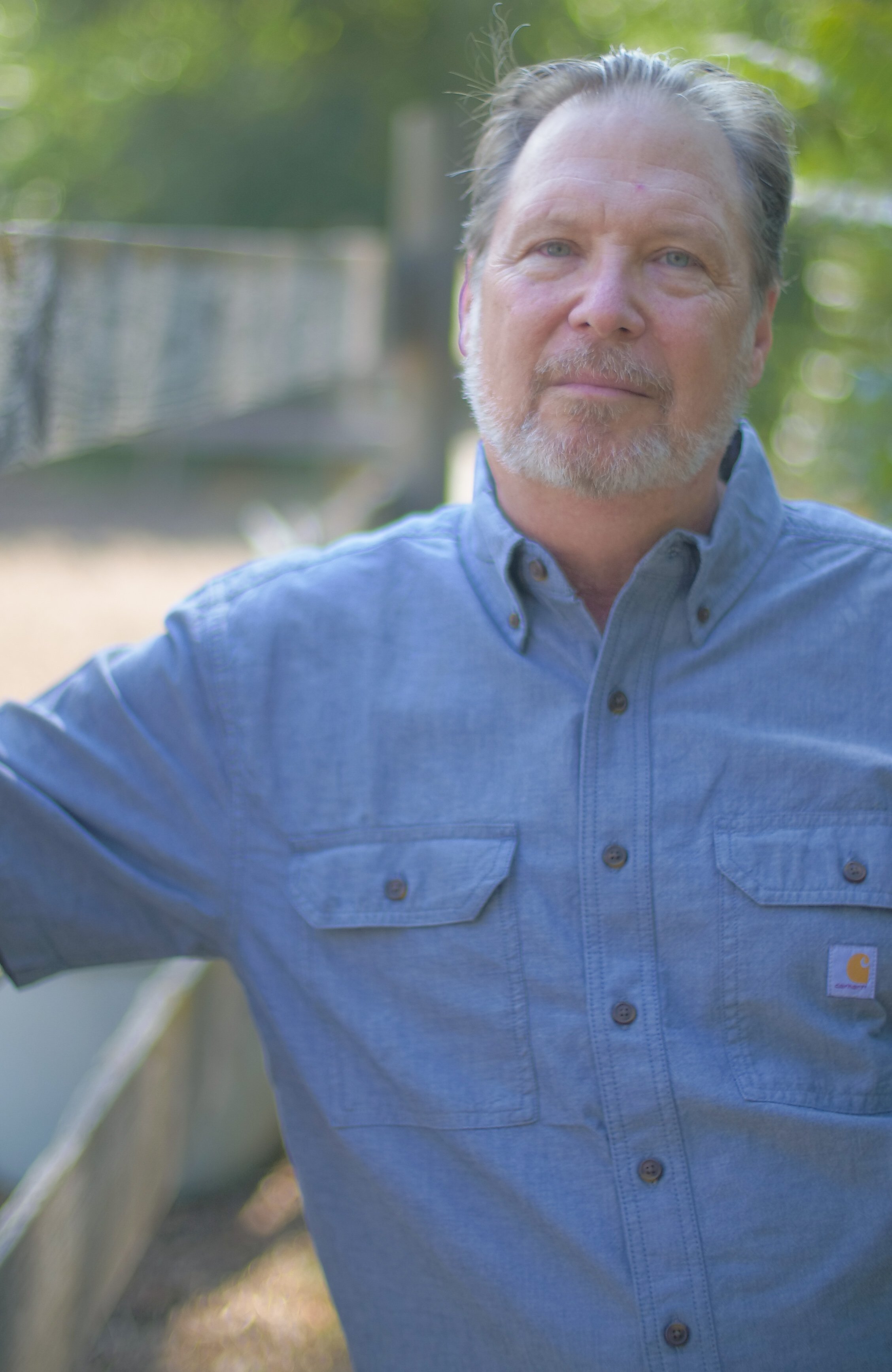 A middle-aged man with gray hair and beard wearing a blue button-up shirt outdoors, with trees and a fence in the background.