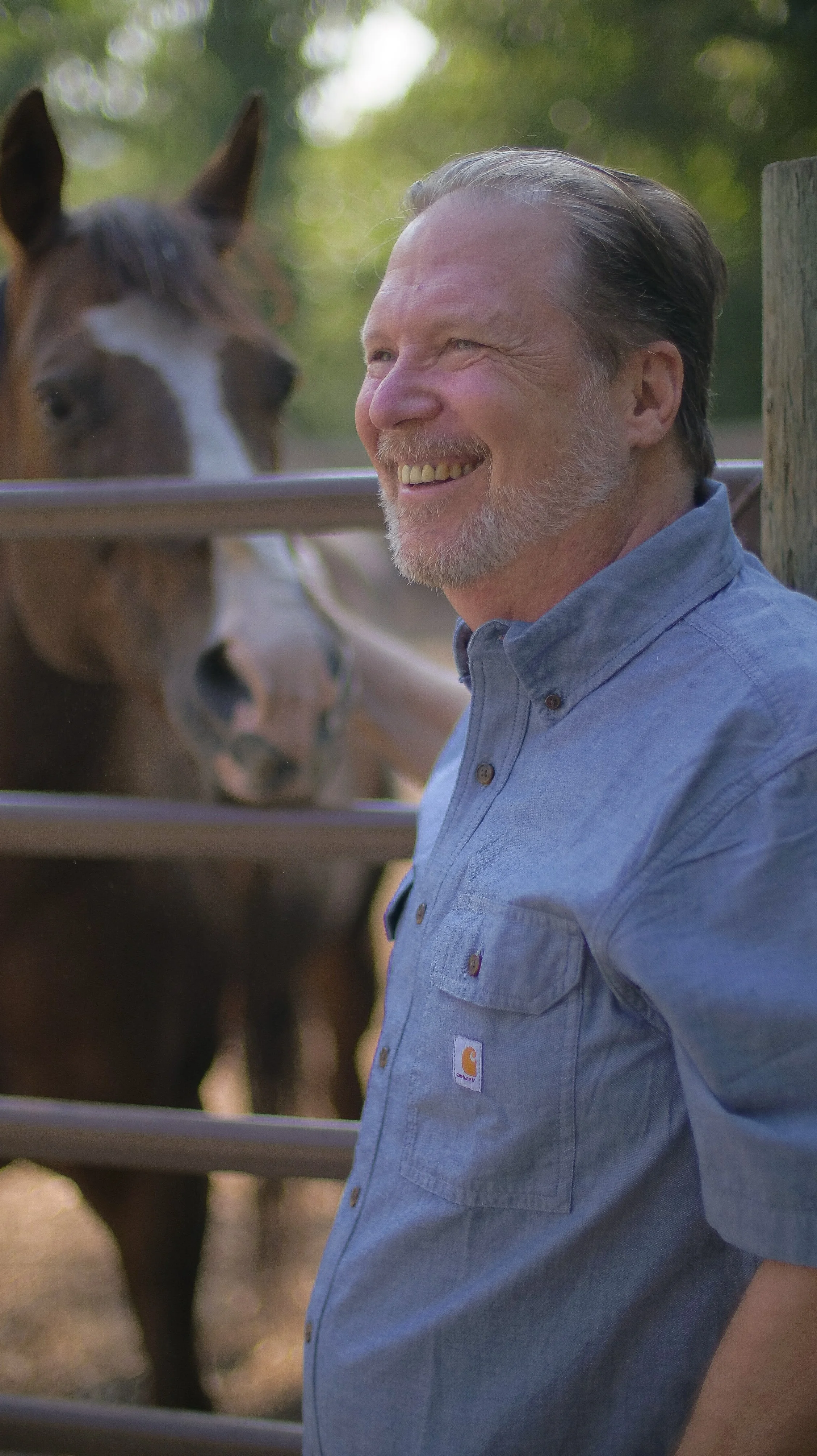 A smiling middle-aged man with a beard and mustache, wearing a blue button-up shirt, standing outdoors near a wooden fence with a horse behind it.