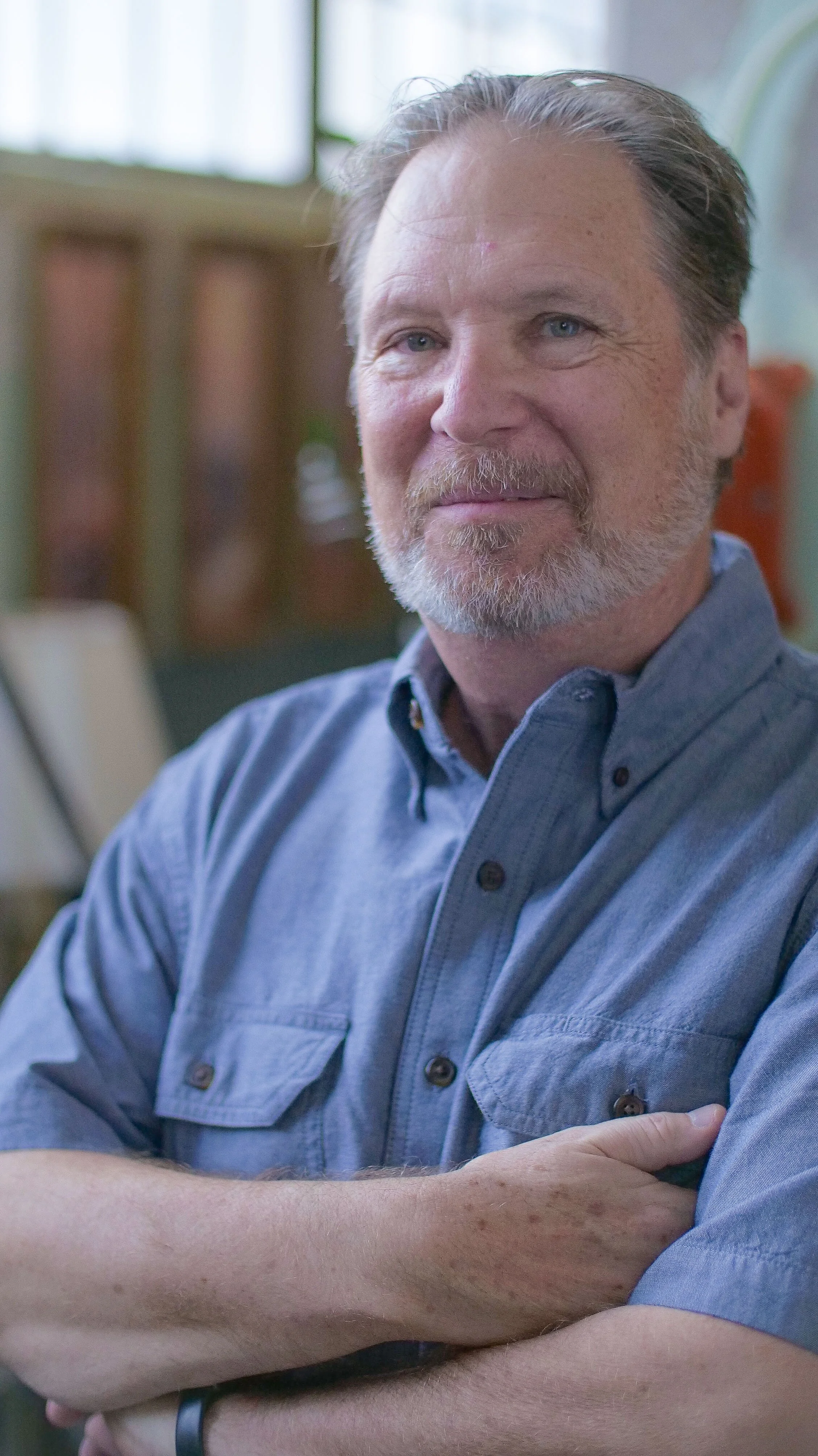 A middle-aged man with light skin, gray hair, a beard, and blue eyes, wearing a blue button-up shirt with folded arms, sitting indoors with blurred background.
