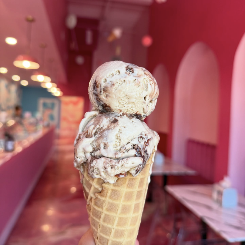 Close-up of a melting ice cream cone with two scoops, one vanilla and chocolate swirl, held in a pink-themed ice cream shop.