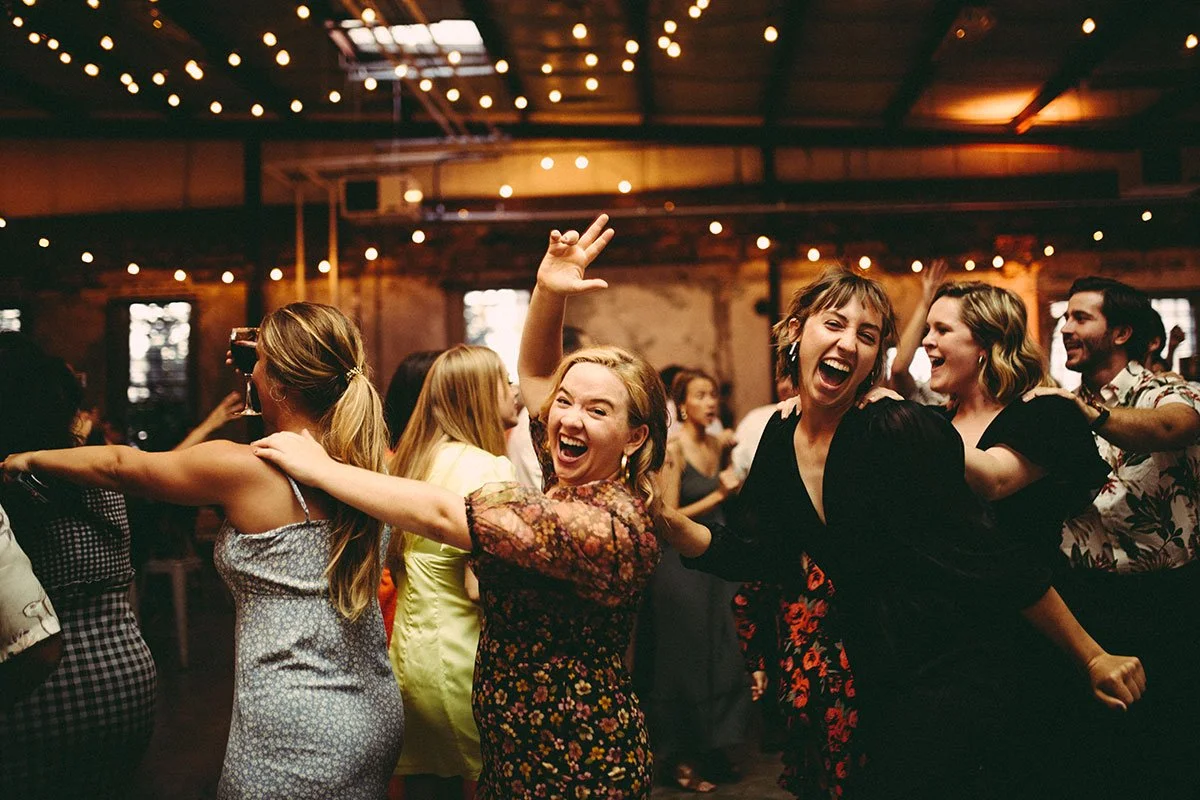 People dancing and celebrating at a lively party with string lights overhead, showing smiling women in the foreground and other guests in the background.