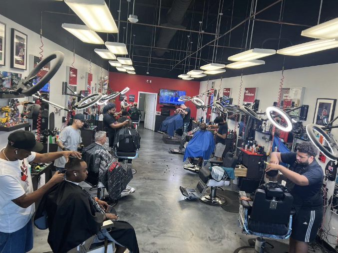 Inside a busy barbershop with multiple barber stations, clients getting haircuts, and barbers working on their clients. There are circular ring lights above each station and various barber supplies visible.