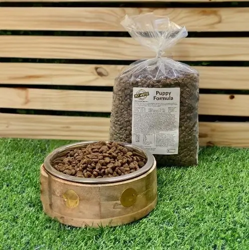 A bowl of dry puppy food and a large bag of puppy food on grass in front of a wooden fence.