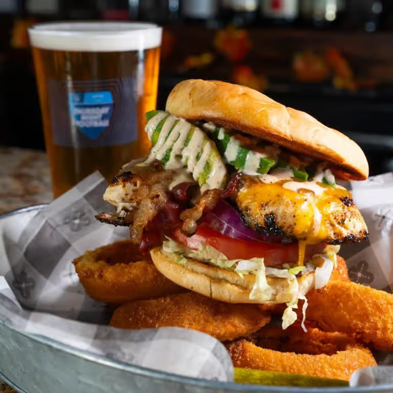 A burger with fried onions, a fried egg, bacon, lettuce, tomato, pickles, and a bun, served with onion rings and a glass of beer in the background.