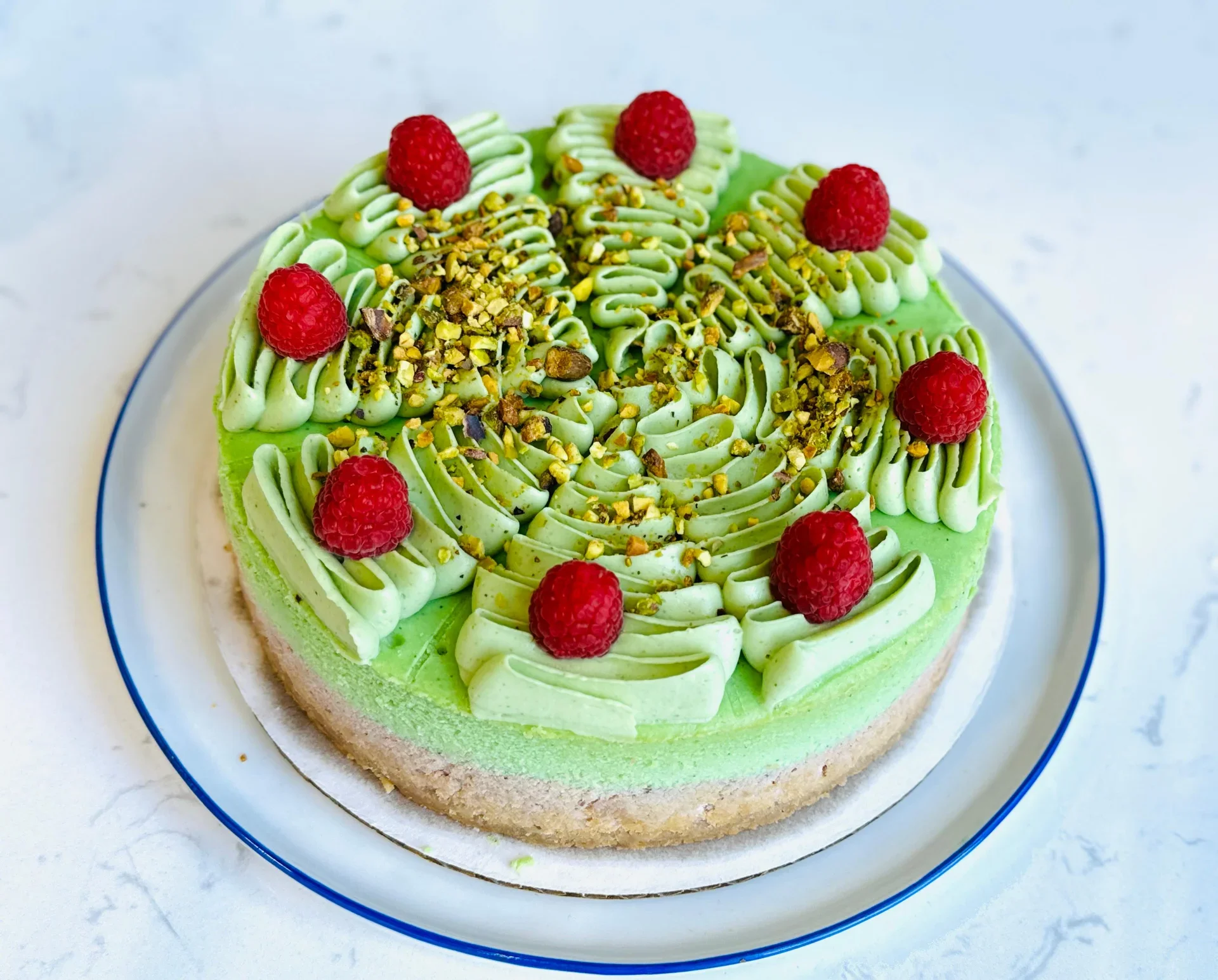 A round cake with green frosting, decorated with red raspberries and chopped nuts on top, placed on a white plate with a blue rim.