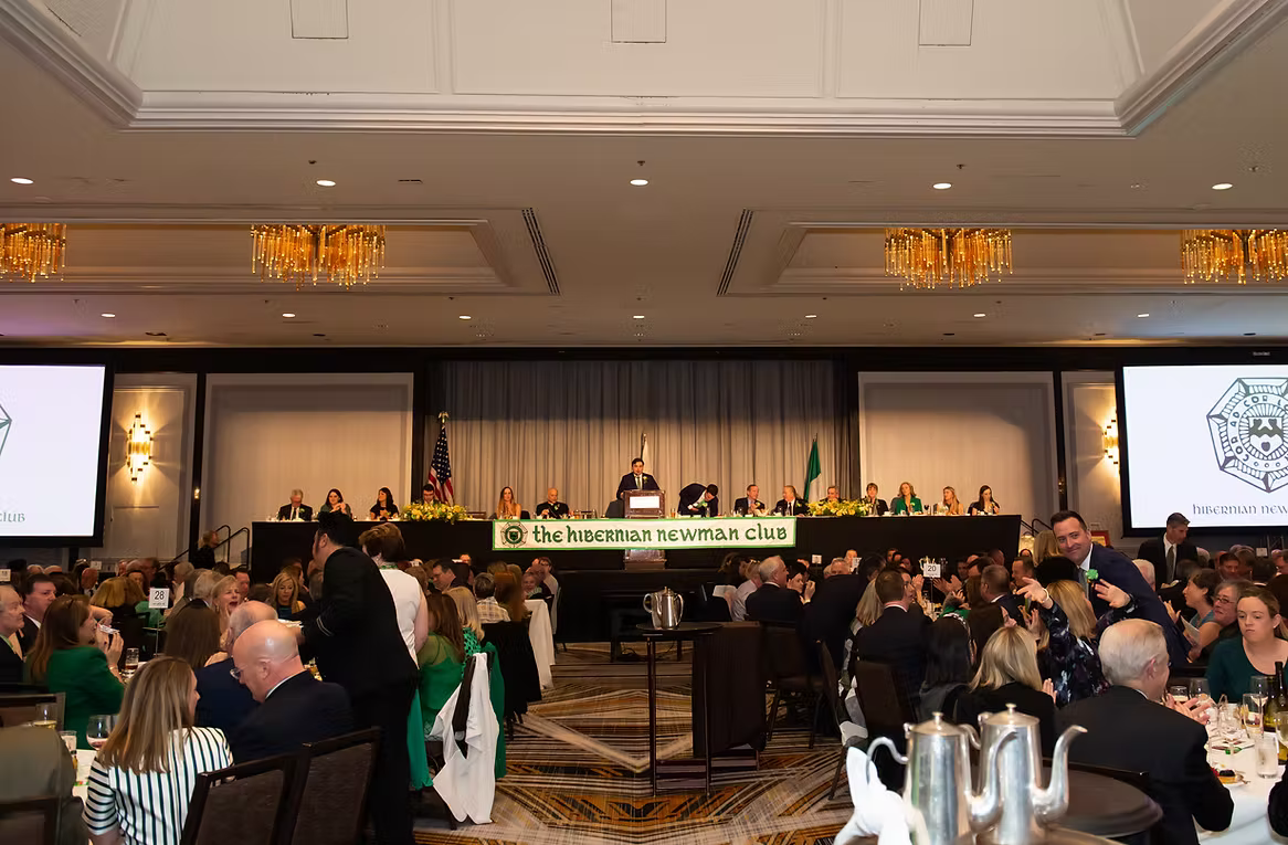 A formal banquet hall filled with people seated at round tables, with a large stage at the front where a speaker is addressing the audience. The stage has a sign reading 'The Hibernian Newman Club' and features two large screens displaying the club's emblem. The hall is decorated with chandeliers and floral arrangements.