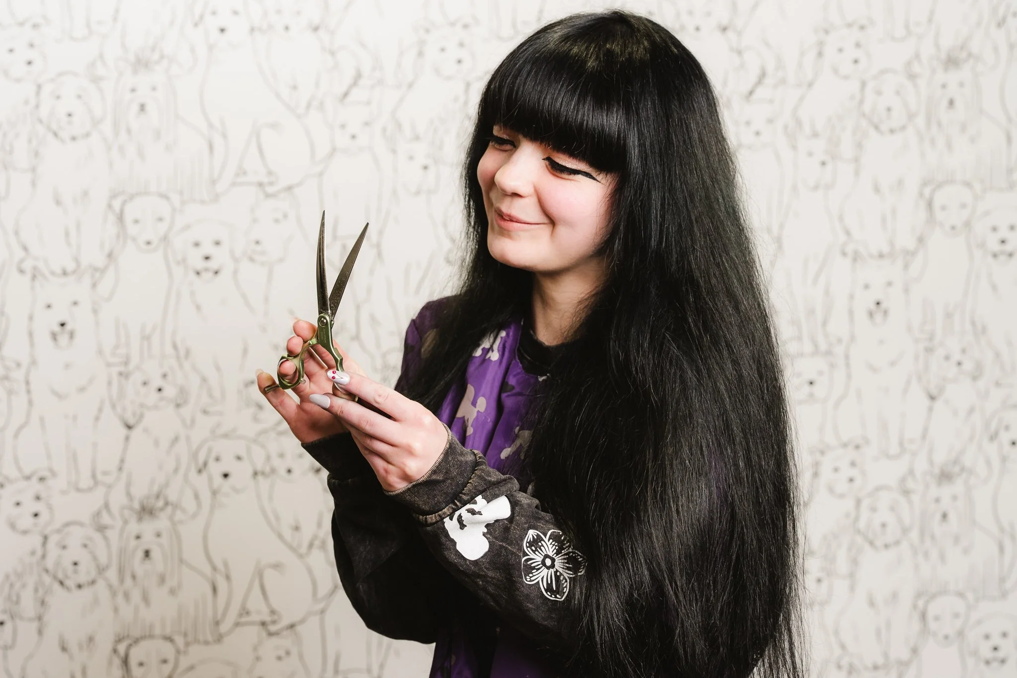 A young woman with long black hair and bangs looks at a pair of scissors she is holding in her hands, standing against a background with a pattern of dogs.