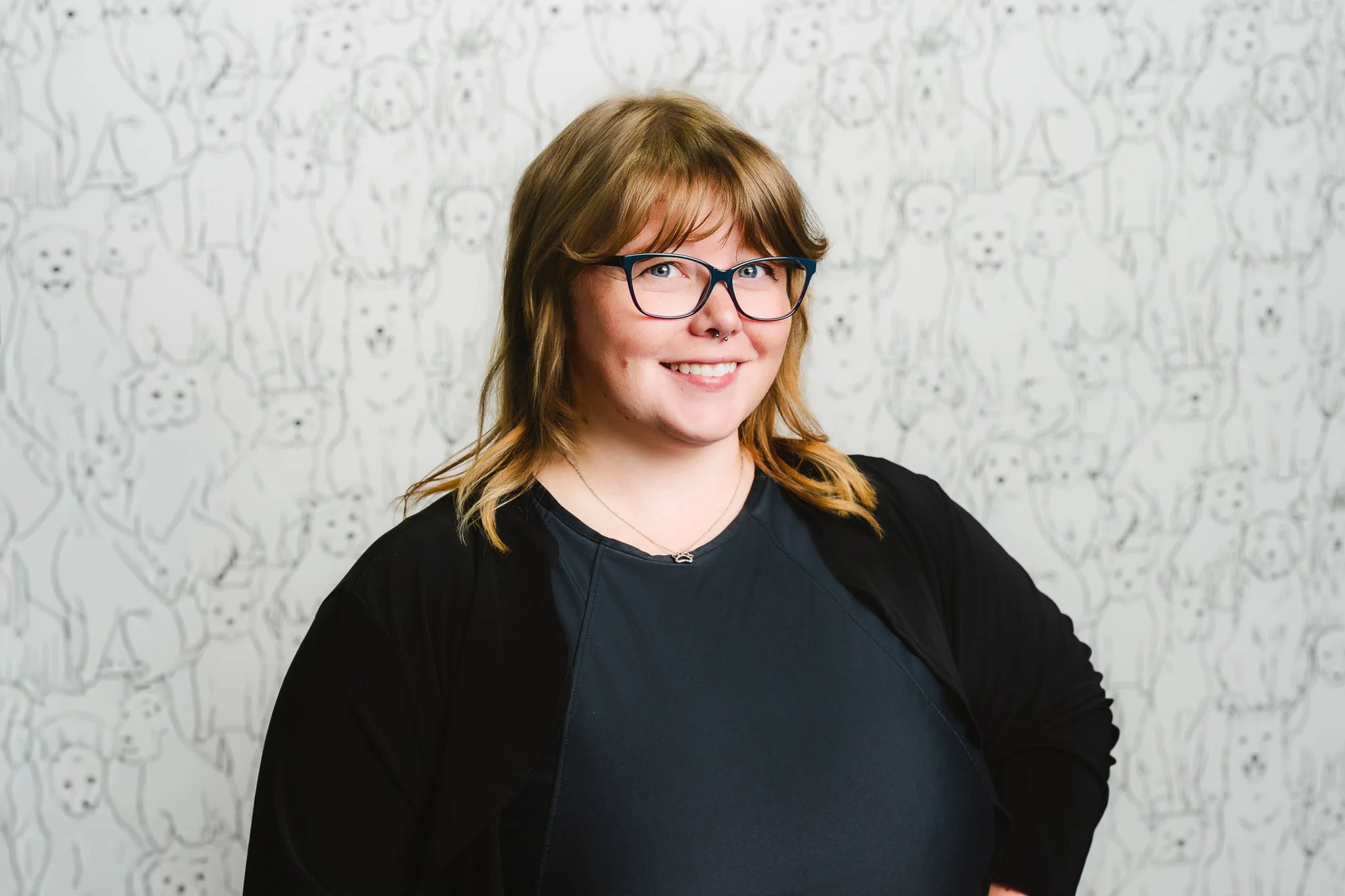 A woman with shoulder-length hair, wearing glasses and a septum piercing, smiling at the camera, standing in front of a white wall with a drawing of multiple bears.