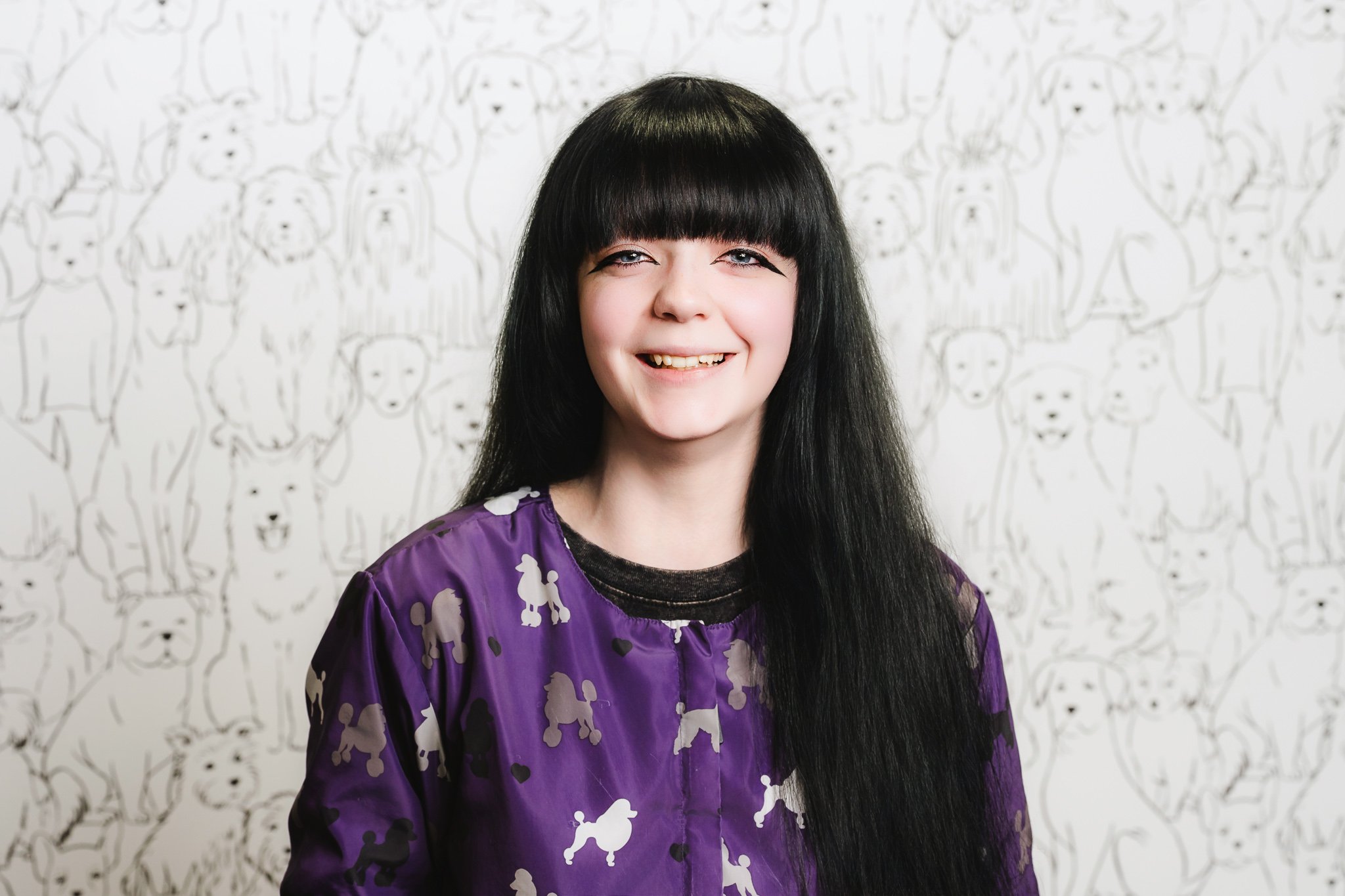A woman with long black hair and bangs, smiling, wearing a purple top with a pattern of white poodles, standing in front of a wall with a repeating dog face pattern.
