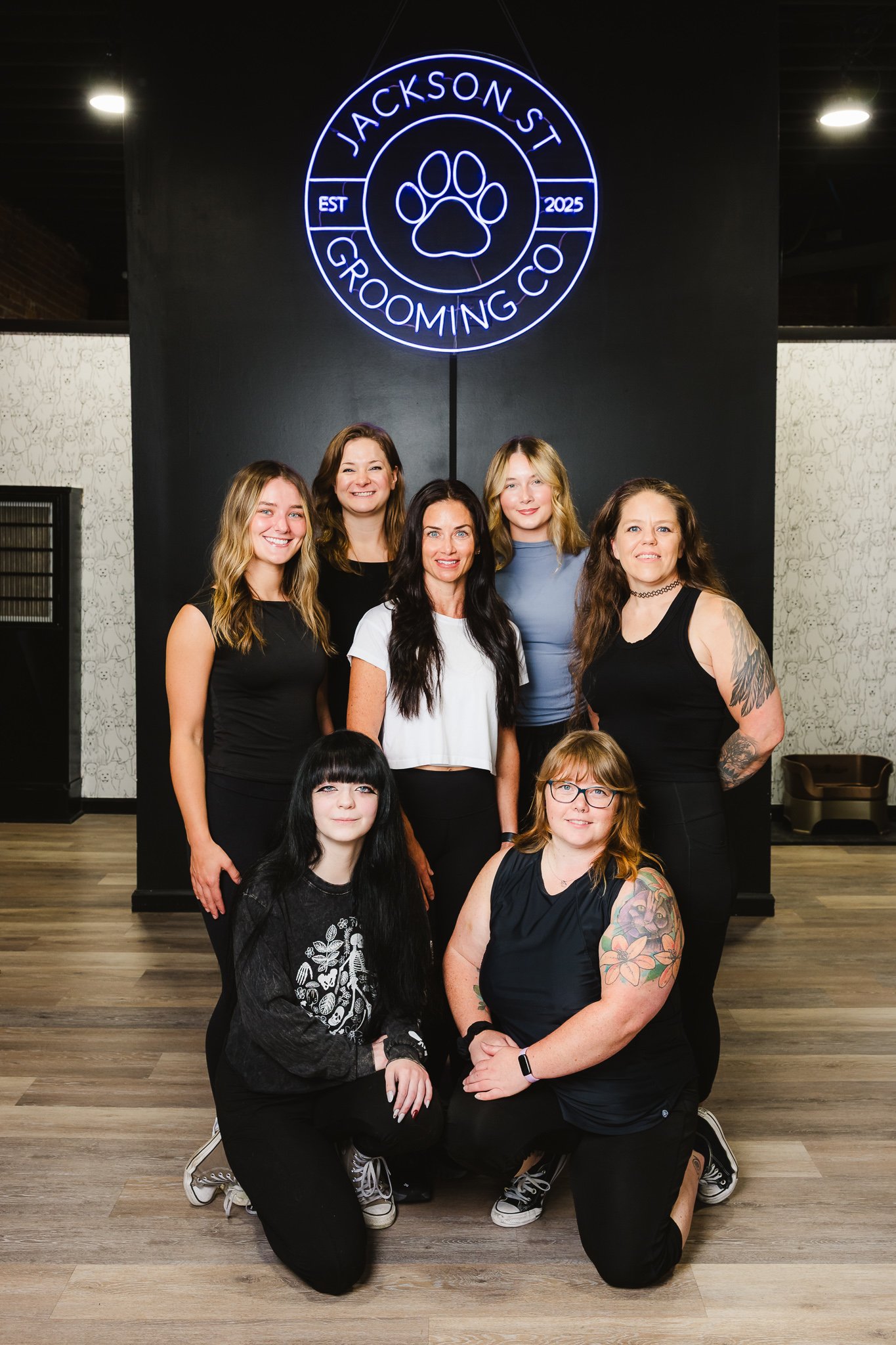 Group of women and a girl posing for a photo inside a grooming center with a neon sign that reads "Jackson St Grooming Co" and features a paw print, established in 2025.