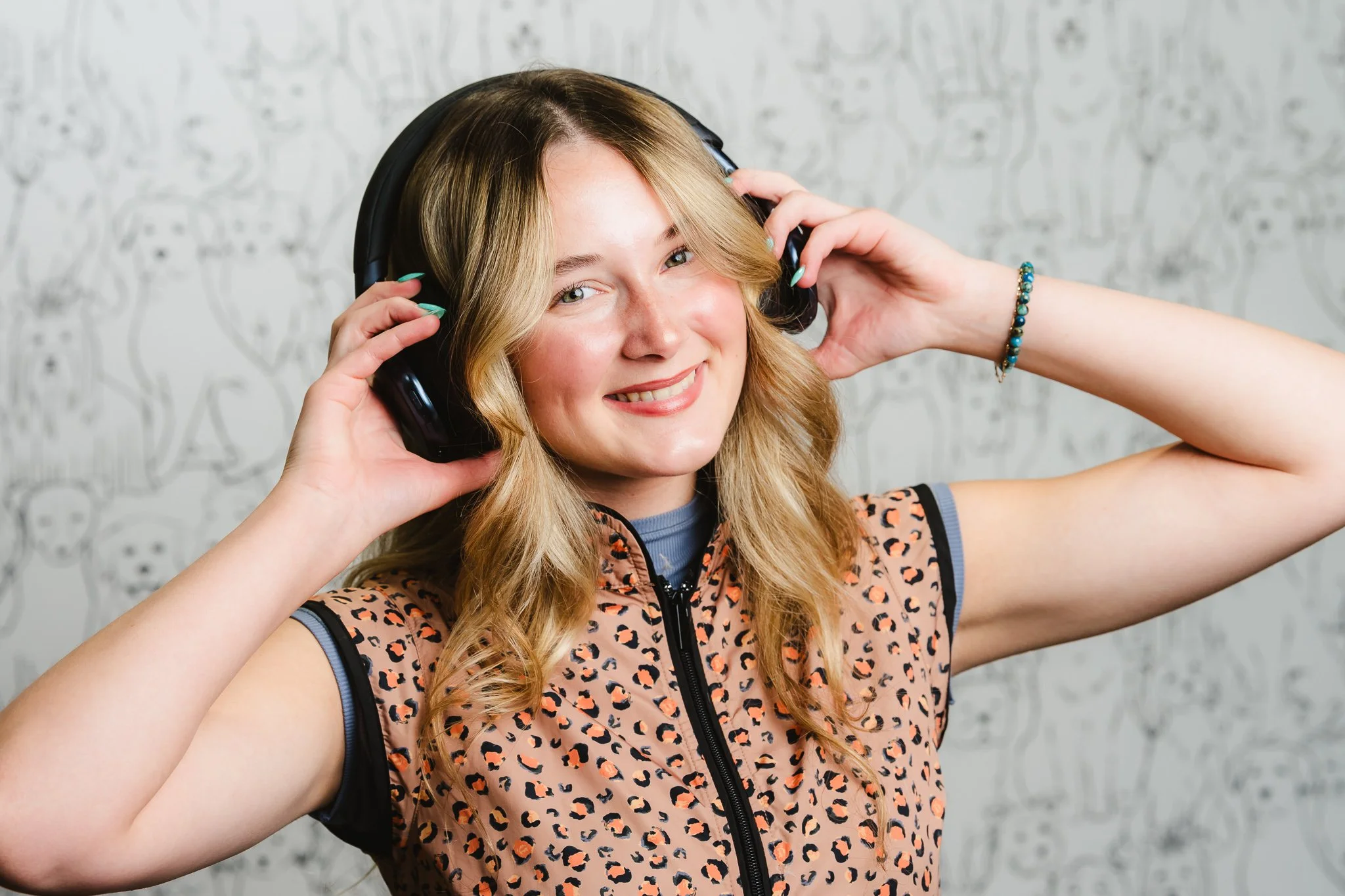 A young woman with long blonde hair smiling, wearing a leopard-print sleeveless top with black trim, holding a large black headphone on her ears, standing in front of a wall with a cartoon dog pattern.