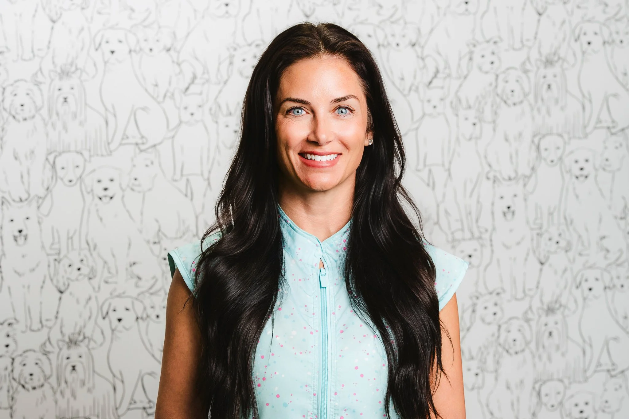 A woman with long dark hair and blue eyes smiling at the camera, standing in front of a white wall with black line drawings of various dog faces.