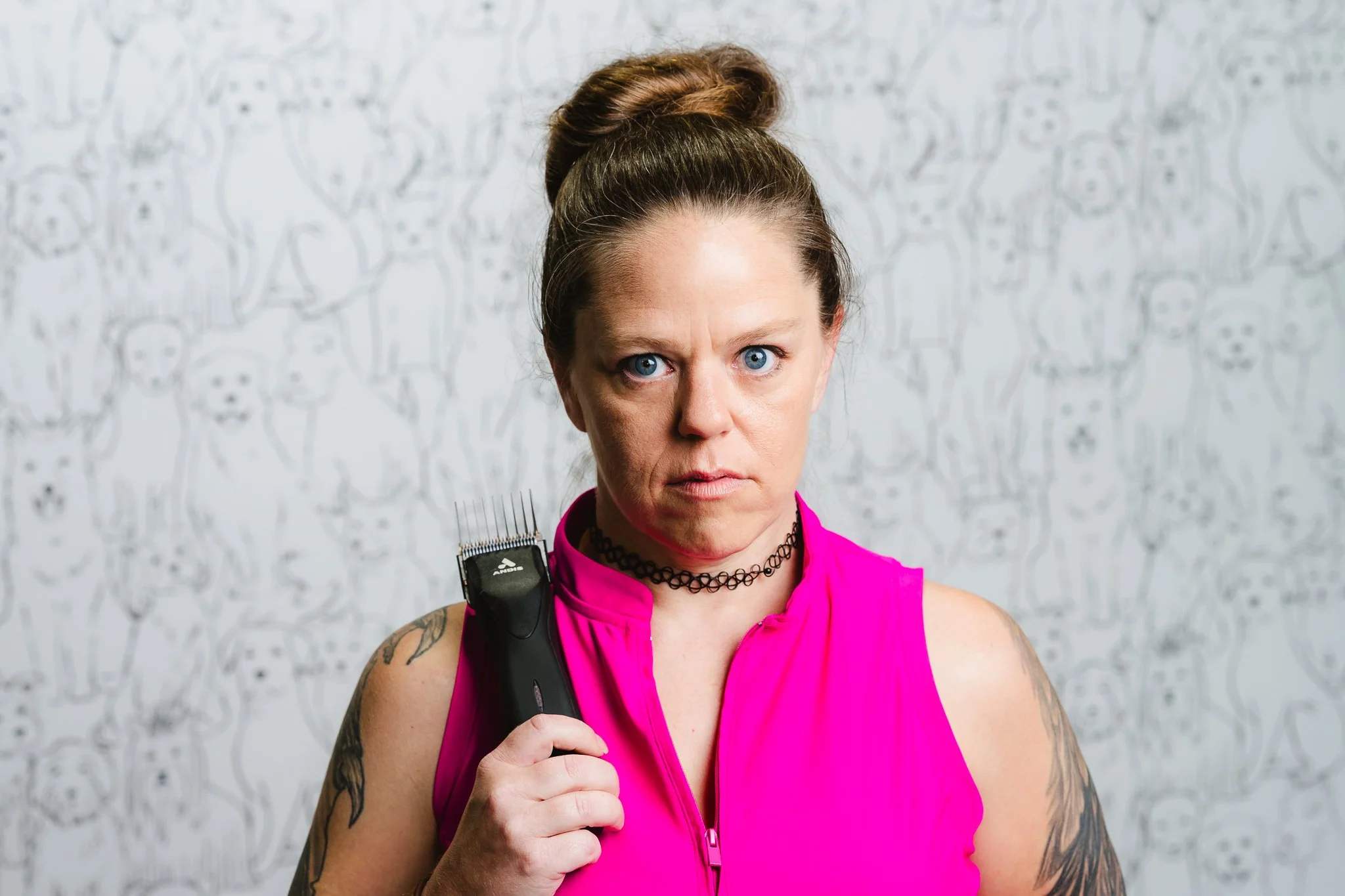 Woman with pink workout top, black choker, holding hair clippers on her shoulder, with tattoos on arms, standing in front of a patterned wall.