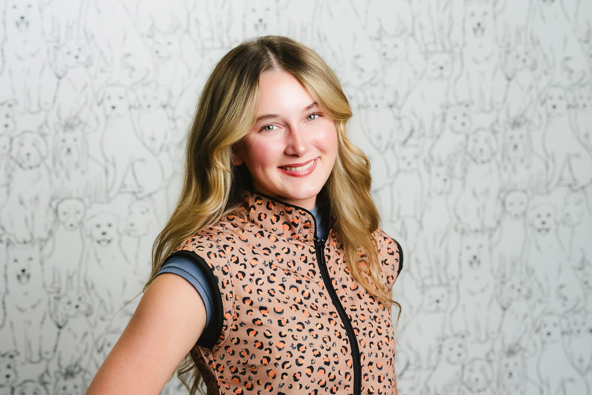 A young woman with long, wavy blonde hair and blue eyes smiling, wearing a pink animal print vest over a blue shirt, standing in front of a wall with a black-and-white dog pattern.