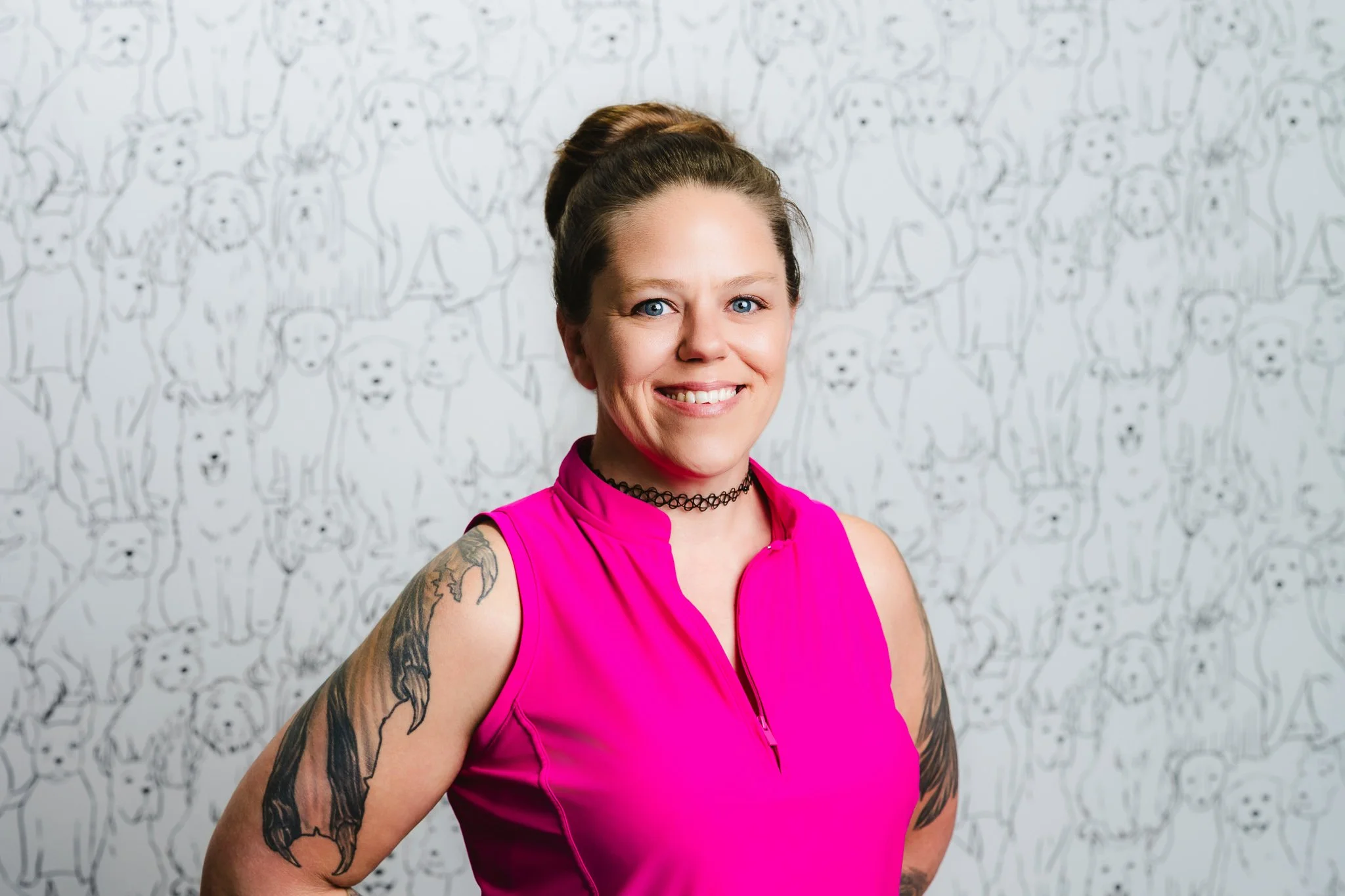 A woman with blue eyes and brown hair in a high bun, smiling and wearing a pink sleeveless top, standing in front of a patterned background of cartoon-style dogs.