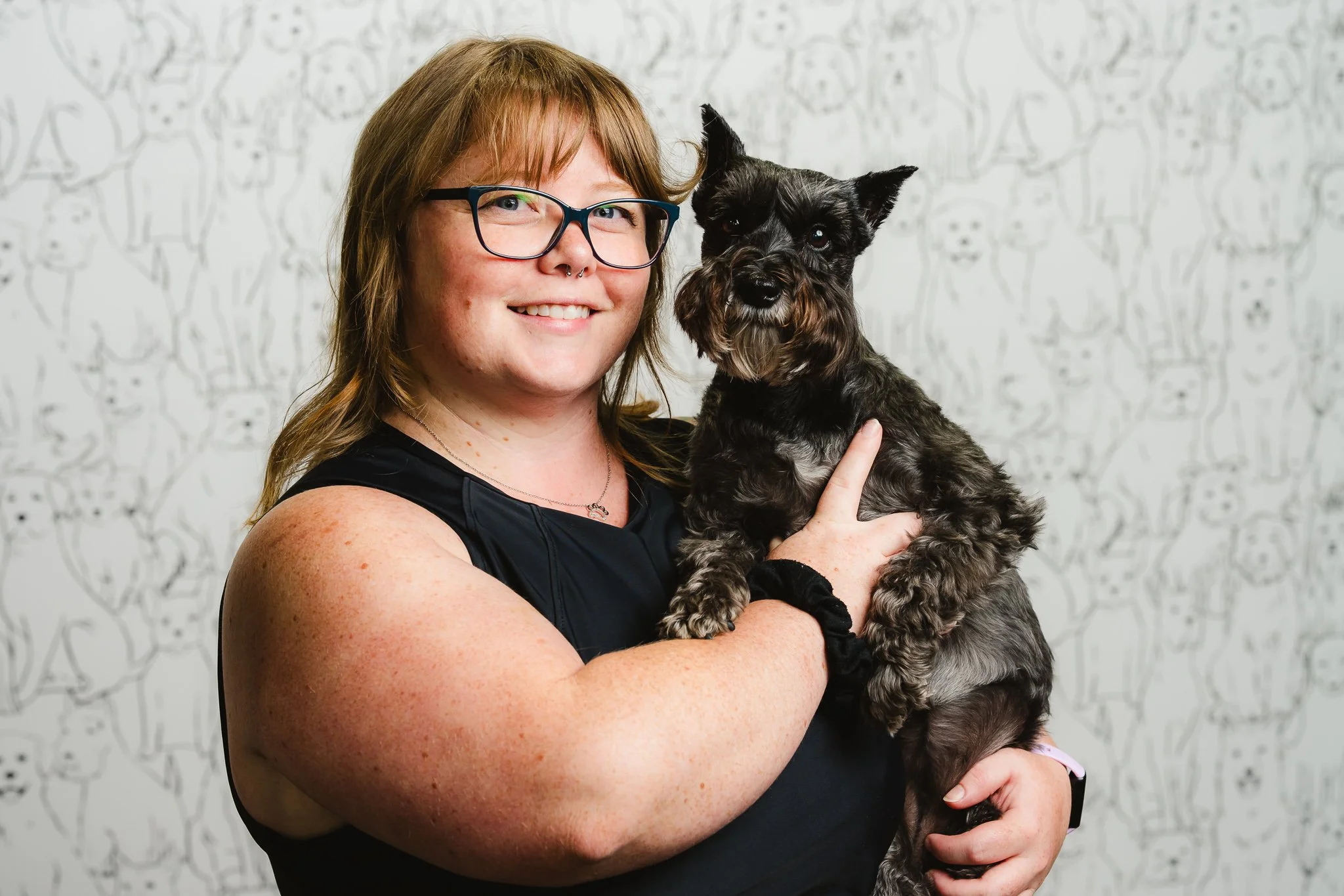 A woman with glasses and short hair holding a small black and gray dog with pointed ears, smiling against a white background with animal illustrations.