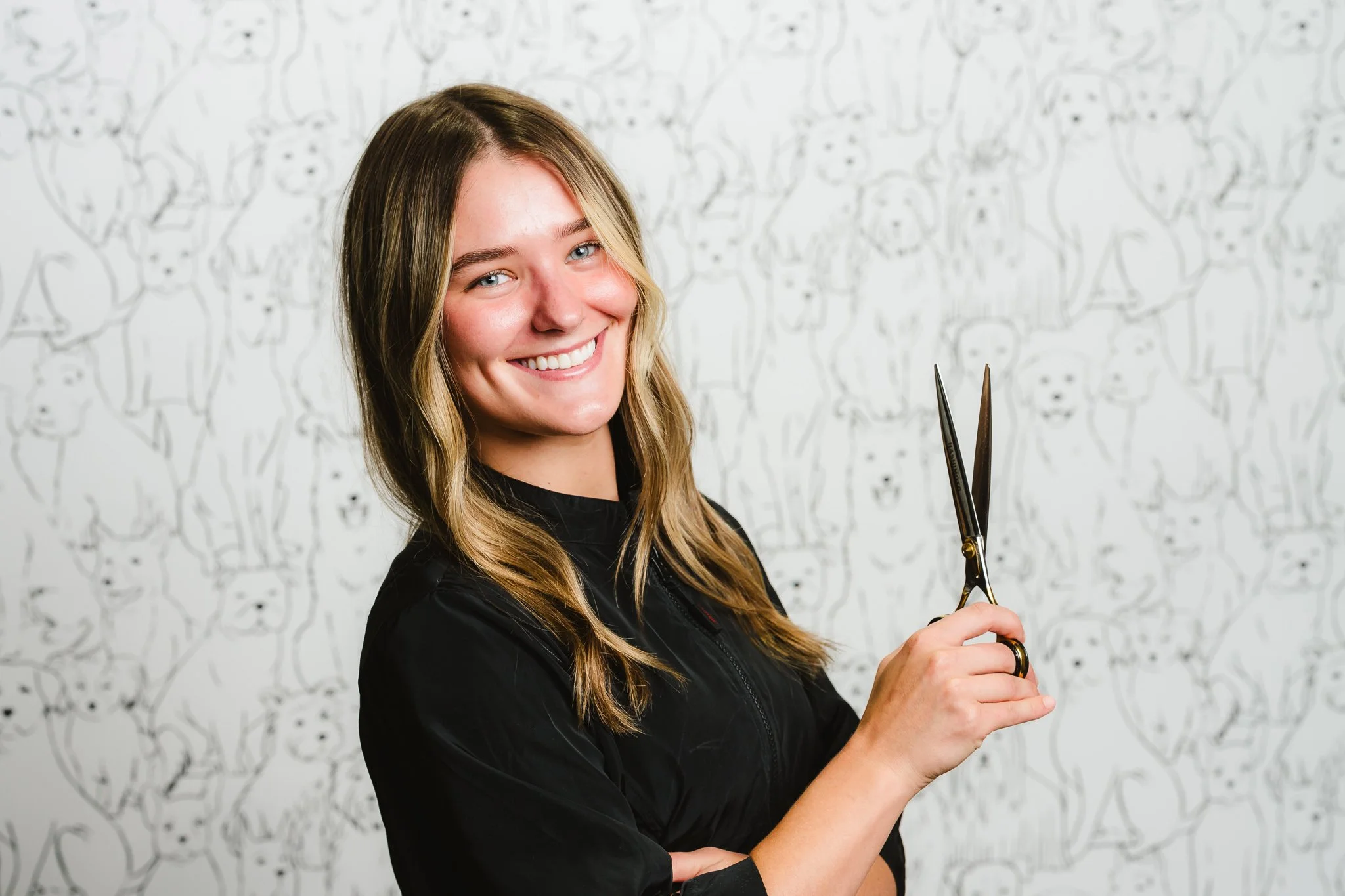 A smiling woman with long blonde hair, wearing a black jacket, holding scissors, in front of a background with a pattern of dog sketches.