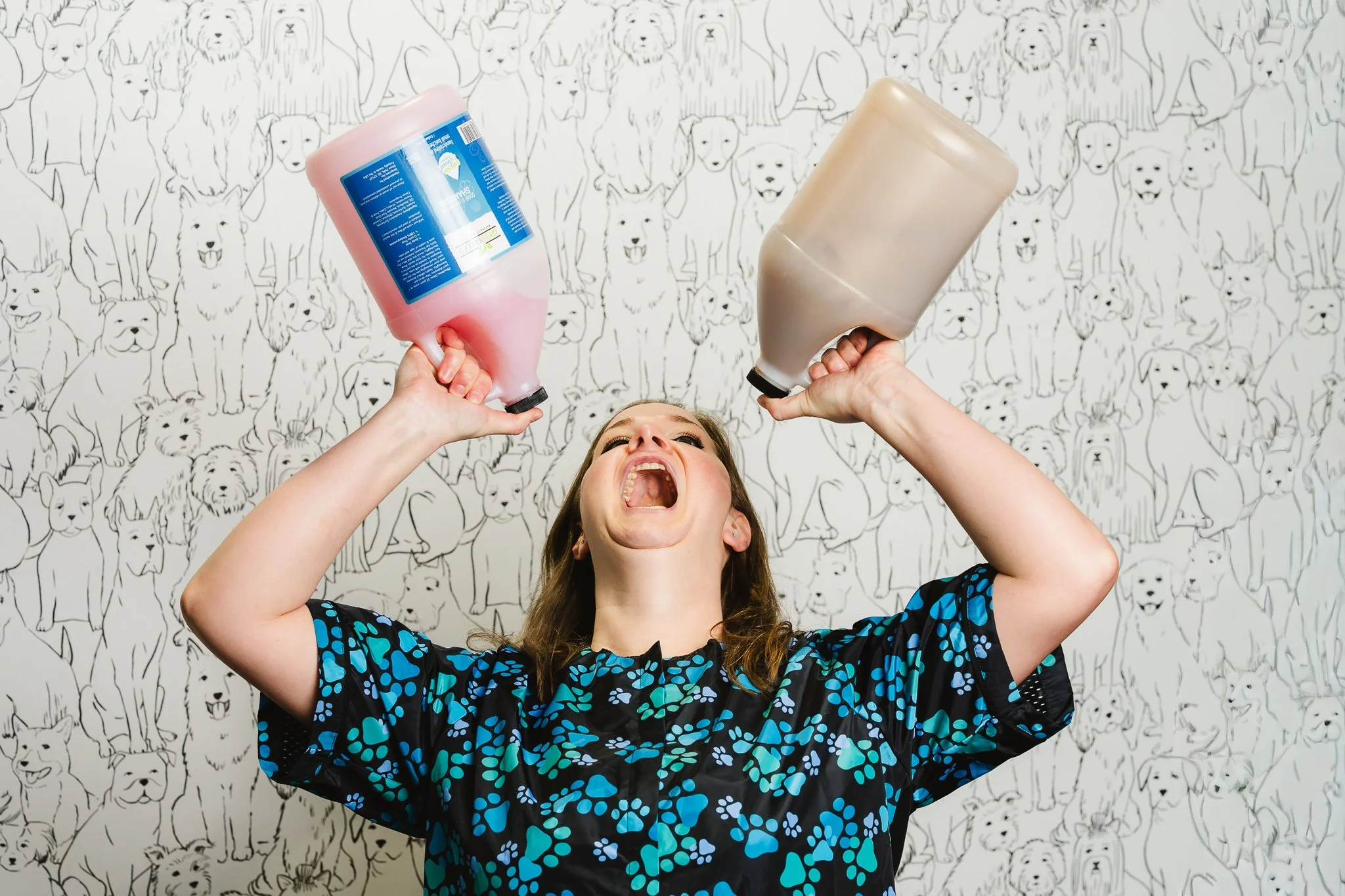Young girl wearing a blue and black patterned shirt, laughing, holding two large bottles of shampoo or conditioner, with her head tilted back and mouth open, in front of a black and white wallpaper with various dog illustrations.