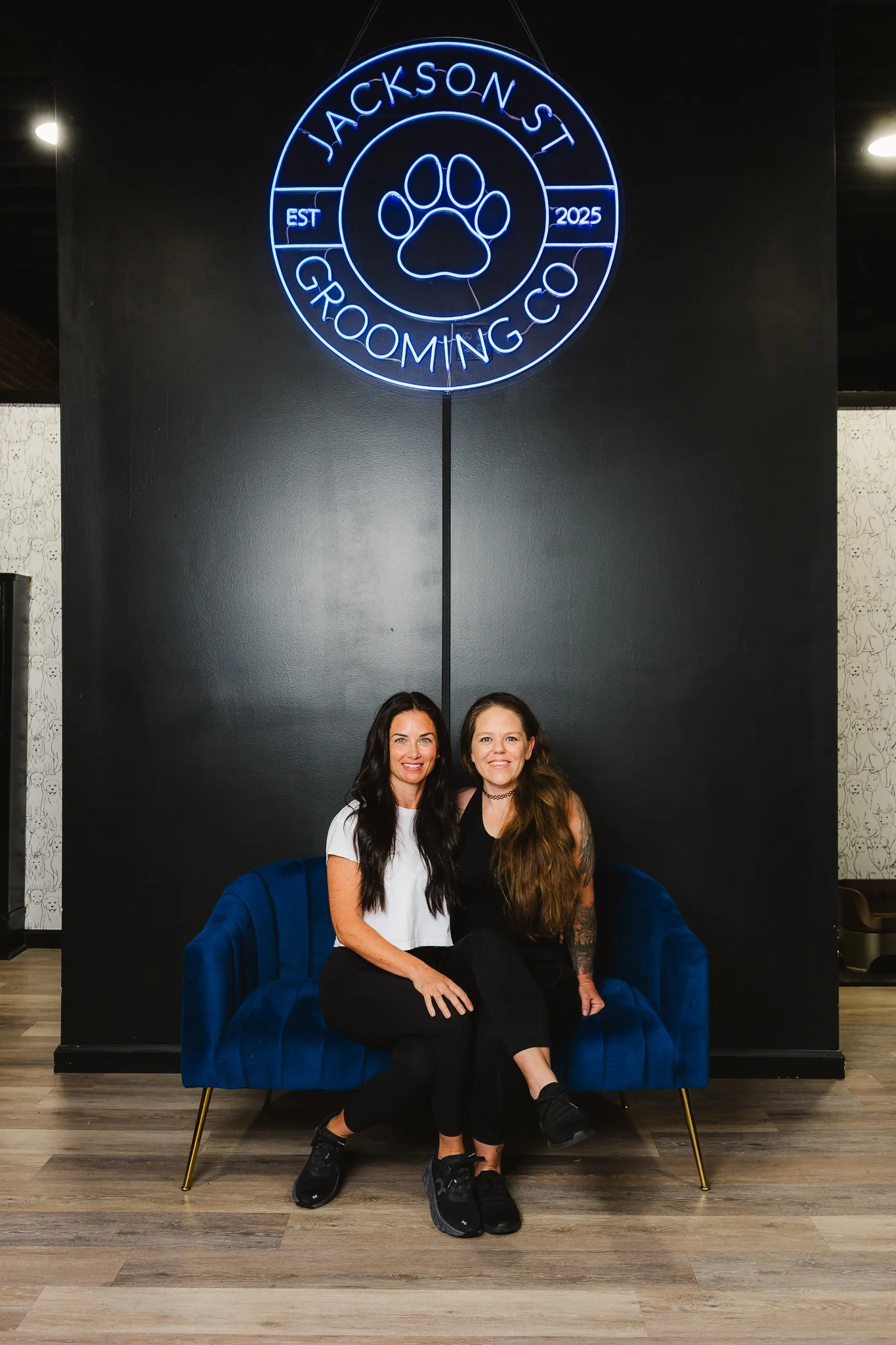Two women sitting on a blue velvet couch in front of a black wall with a neon sign that reads 'Jackson's Grooming Co. EST 2025' and has a paw print design in the middle.