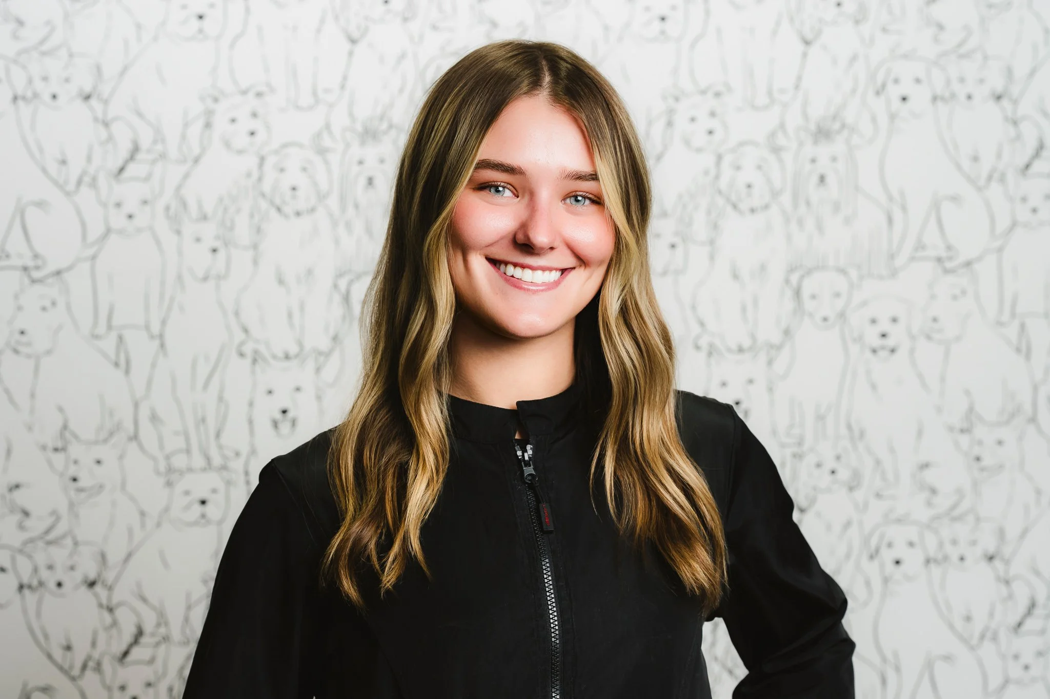 Smiling young woman with long wavy blonde hair standing in front of a white wall with a pattern of hand-drawn dog faces.