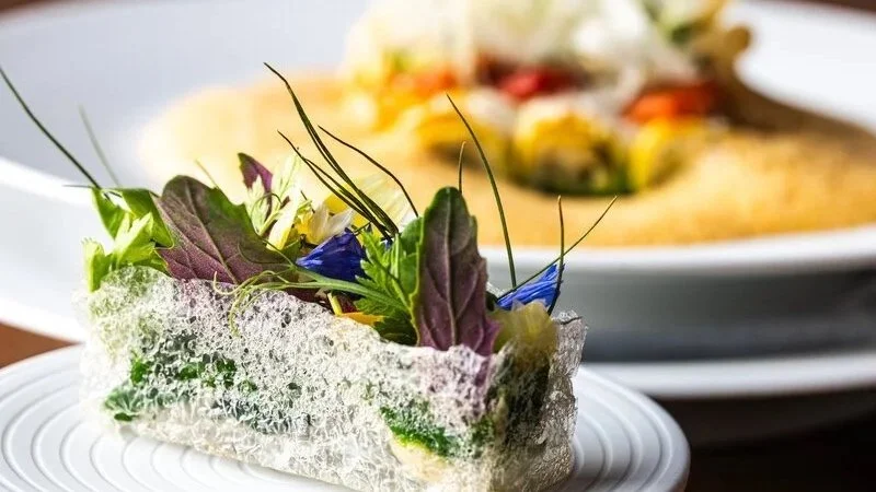 Feines, kreatives Gericht mit einer knusprigen, transparenten Hülle, gefüllt mit grünem Salat, Kräutern und essbaren Blumen, auf einem weißen Teller, mit weiteren ähnlichen Speisen im Hintergrund.