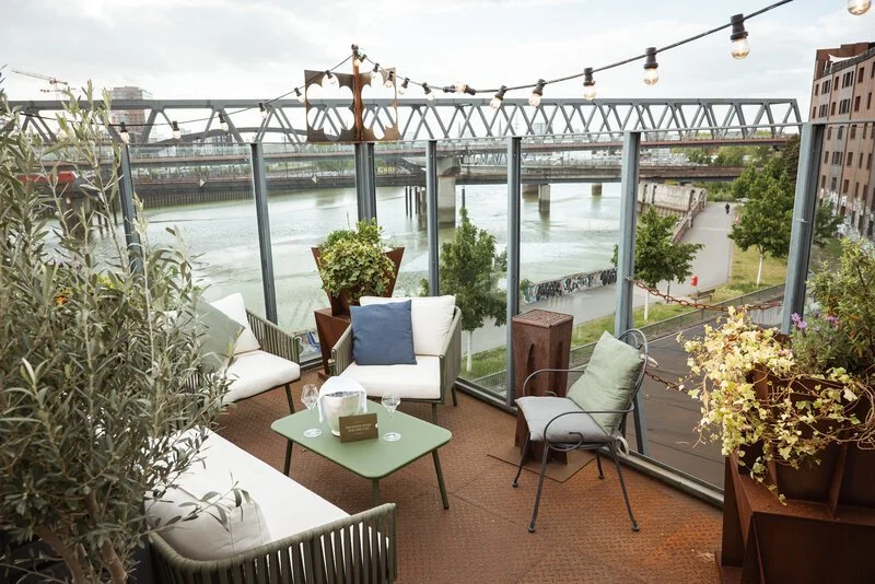 Gemütlicher Balkon mit Sitzmöbeln, Pflanzen und Lichterketten, Blick auf den Fluss und die Brücke im Stadtzentrum.