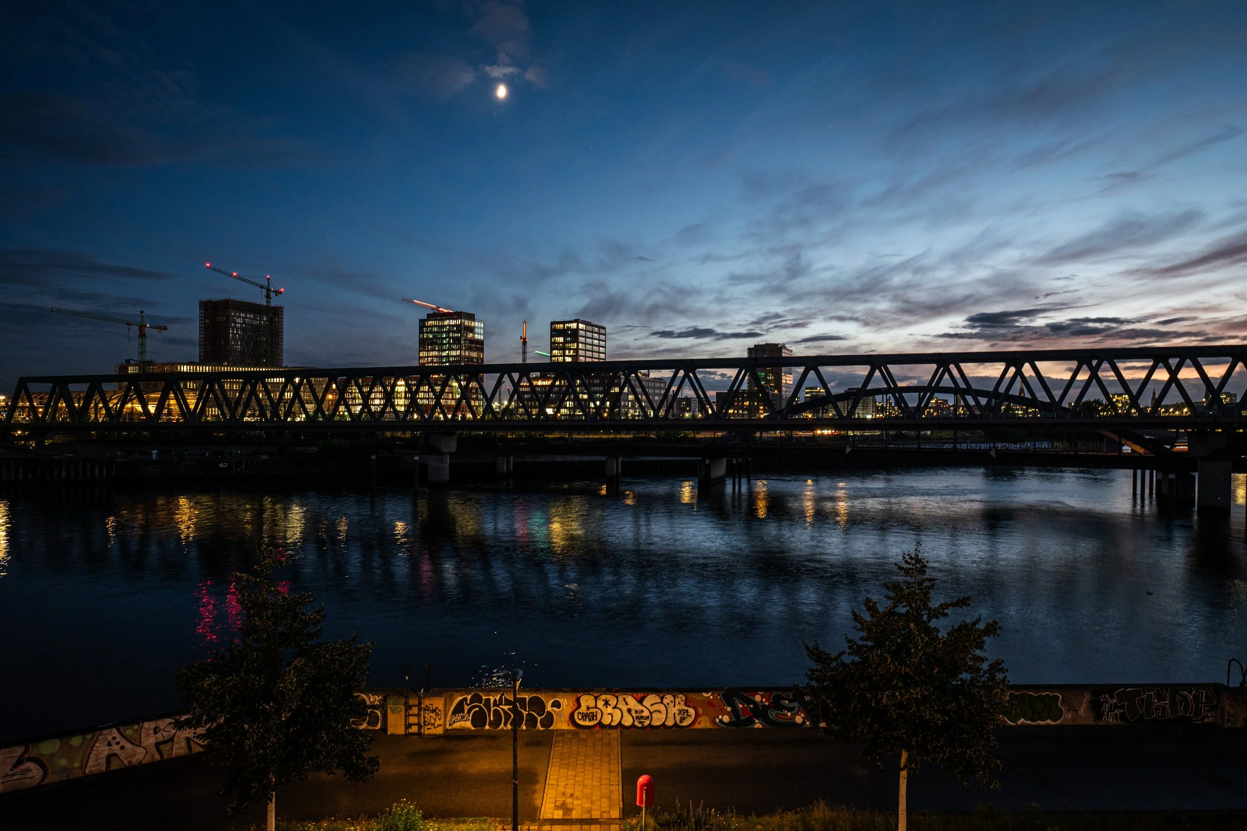 Stadt bei Nacht mit beleuchteten Gebäuden, Brücke über den Fluss, Graffiti an einer Mauer im Vordergrund, bewölkter Himmel mit Mond