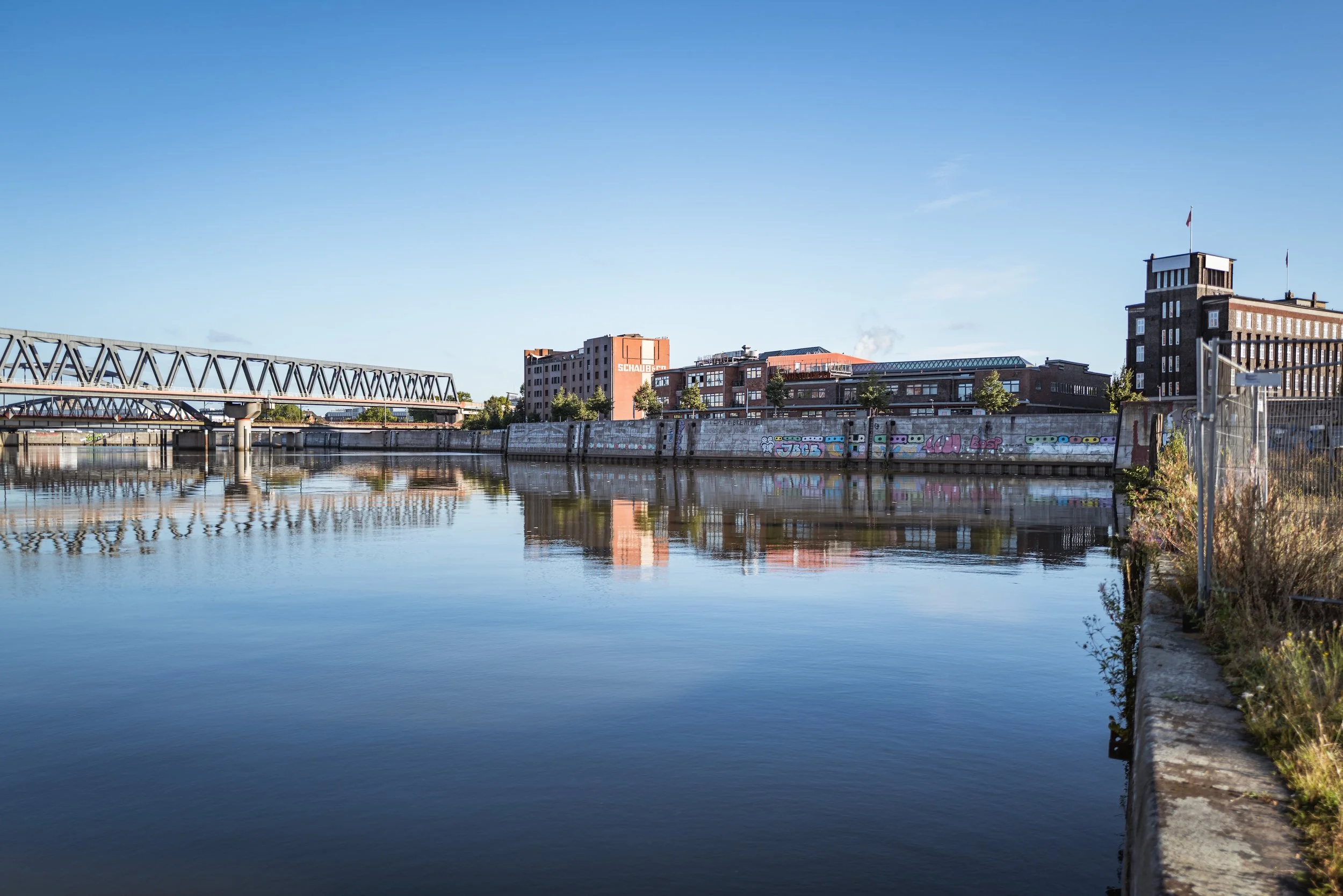 Stadthafen mit Gebäuden, Brücke und Graffiti an der Ufermauer, Wasser spiegelt die Szenerie wider, blauer Himmel, einige Wolken.