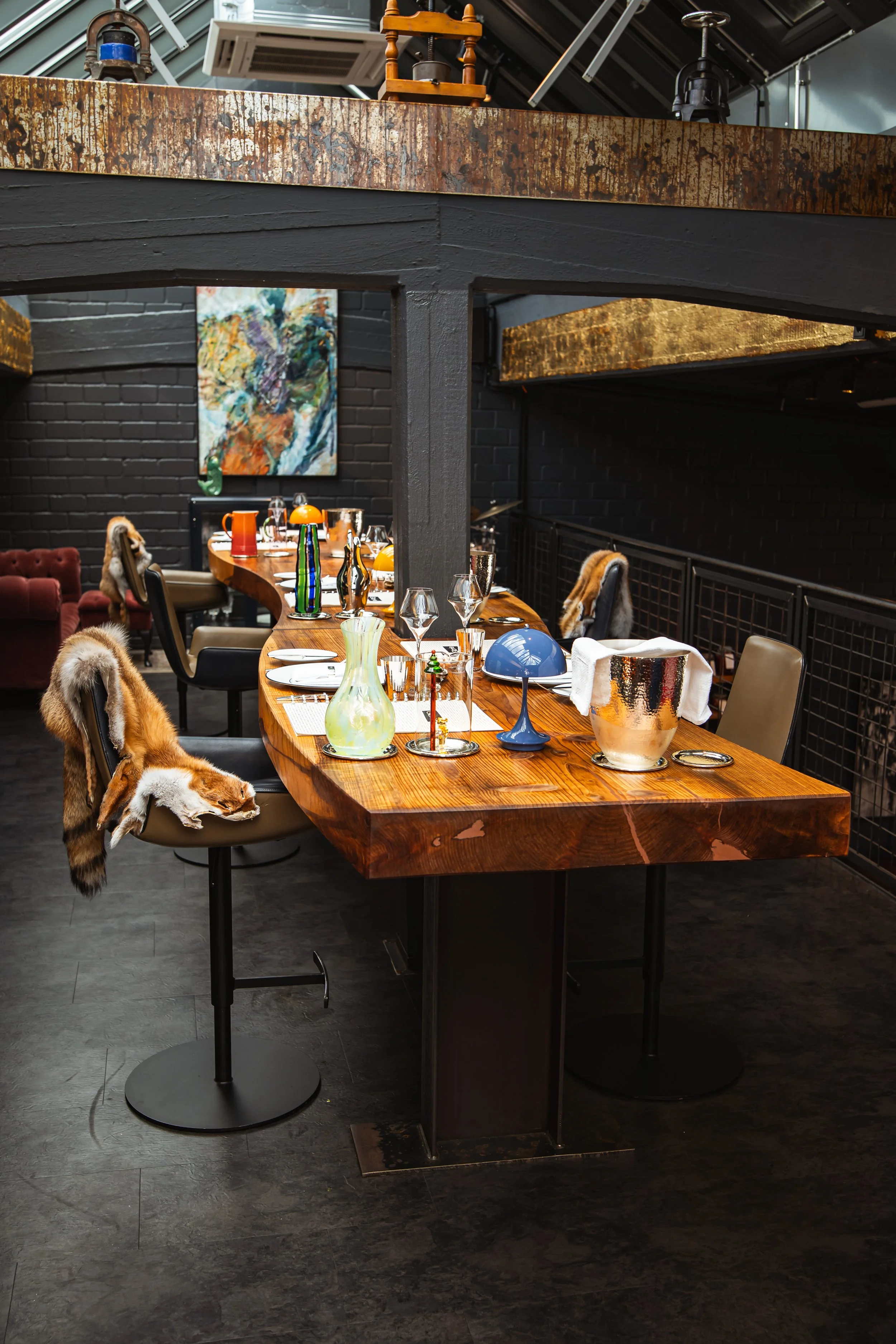 Ein schön gedeckter Esstisch mit Gläsern, Vasen und Besteck in einem modernen Restaurant, mit Schwarz gemauerten Wänden und abstrakter Kunst an der Wand. Im Hintergrund sind einige Stühle mit Felldecken.