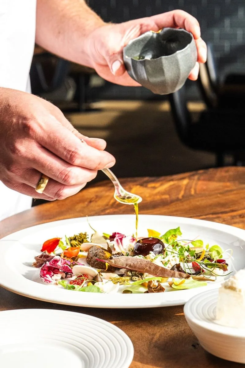 Ein Koch garniert einen bunten Salat auf einem weißen Teller mit Öl oder Dressing. Im Bild sind Hände, ein Löffel und eine Schüssel mit weiterer Zubereitung zu sehen.